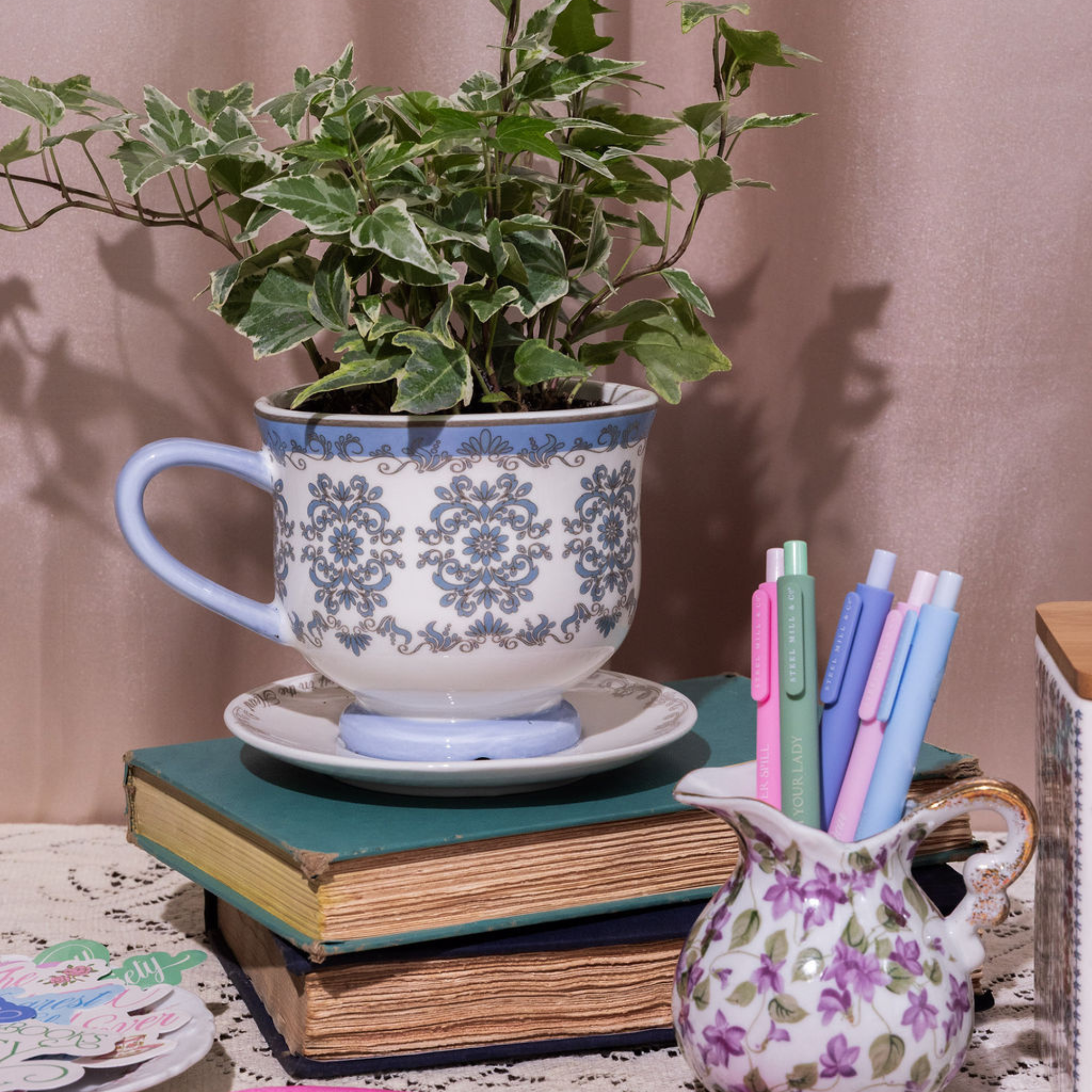 Decorative setup with a plant in a teacup, books, and pens on a table.