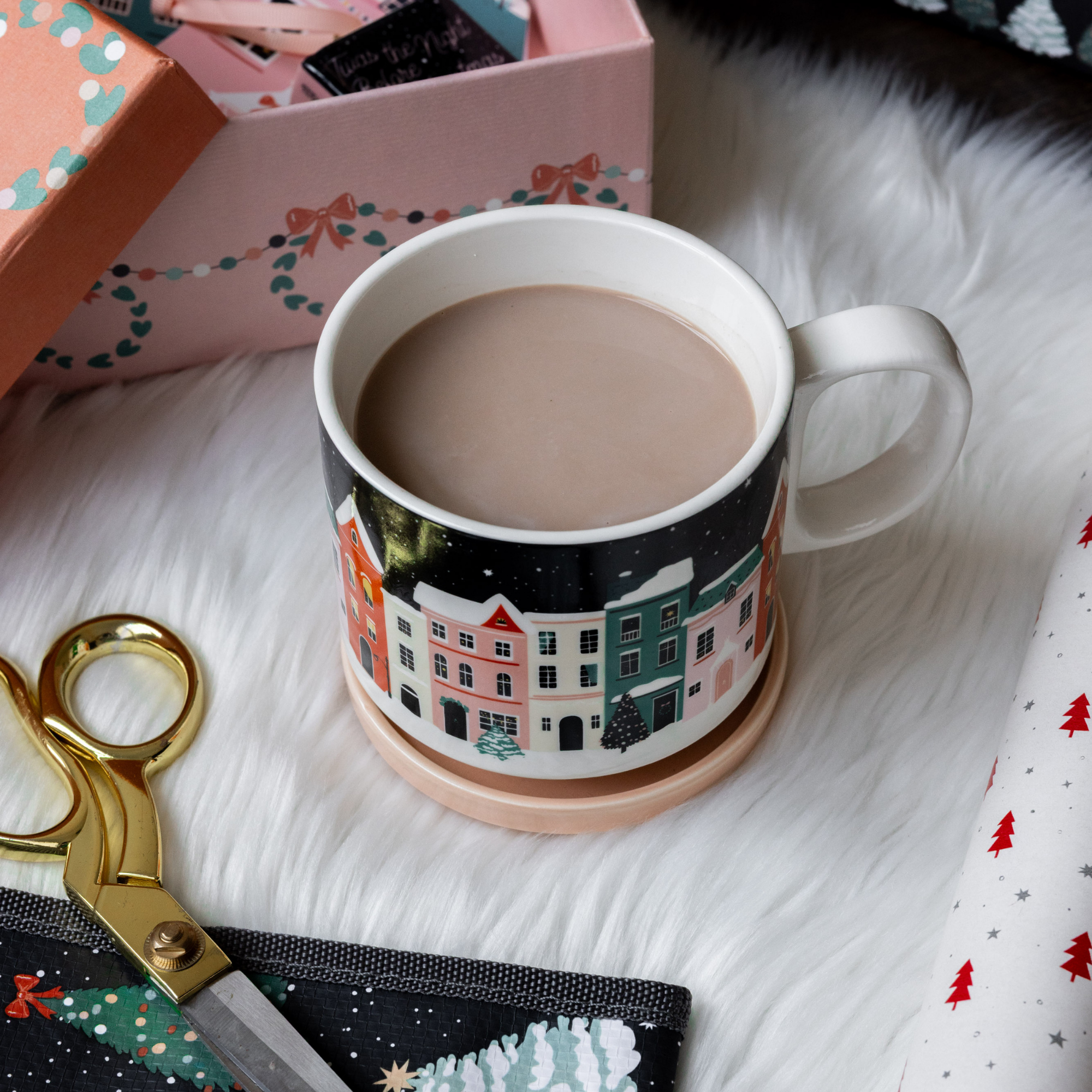 Mug with a decorative design of buildings, filled with a hot beverage, on a white surface with a pair of scissors and a gift box in the background.