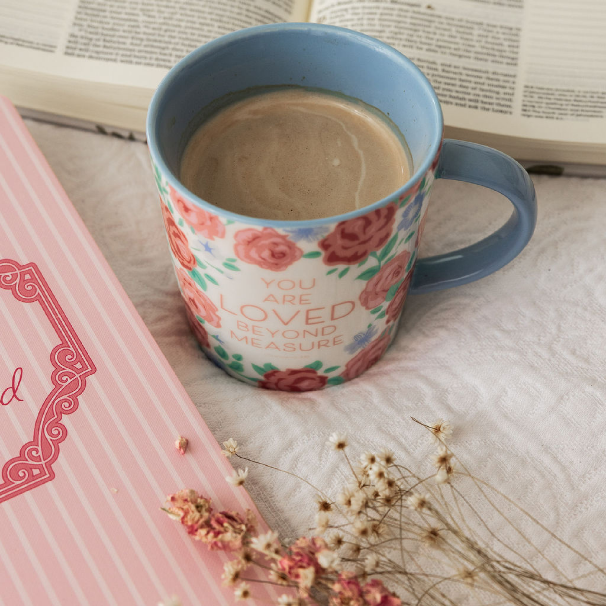 Floral mug with 'You are loved beyond measure' text on a surface with a book and dried flowers.