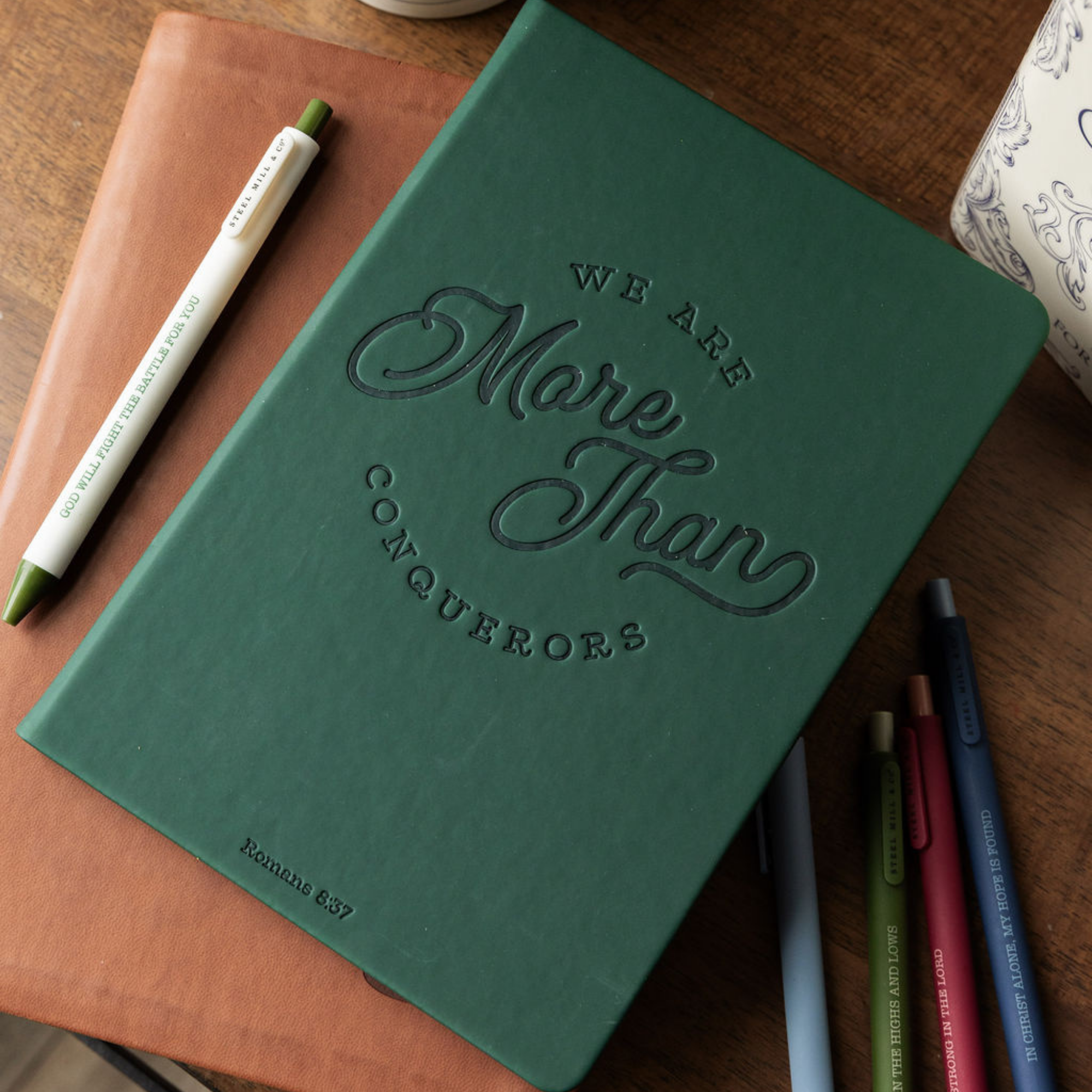 Green notebook with 'We Are More Than Conquerors' text on a wooden surface with pens.