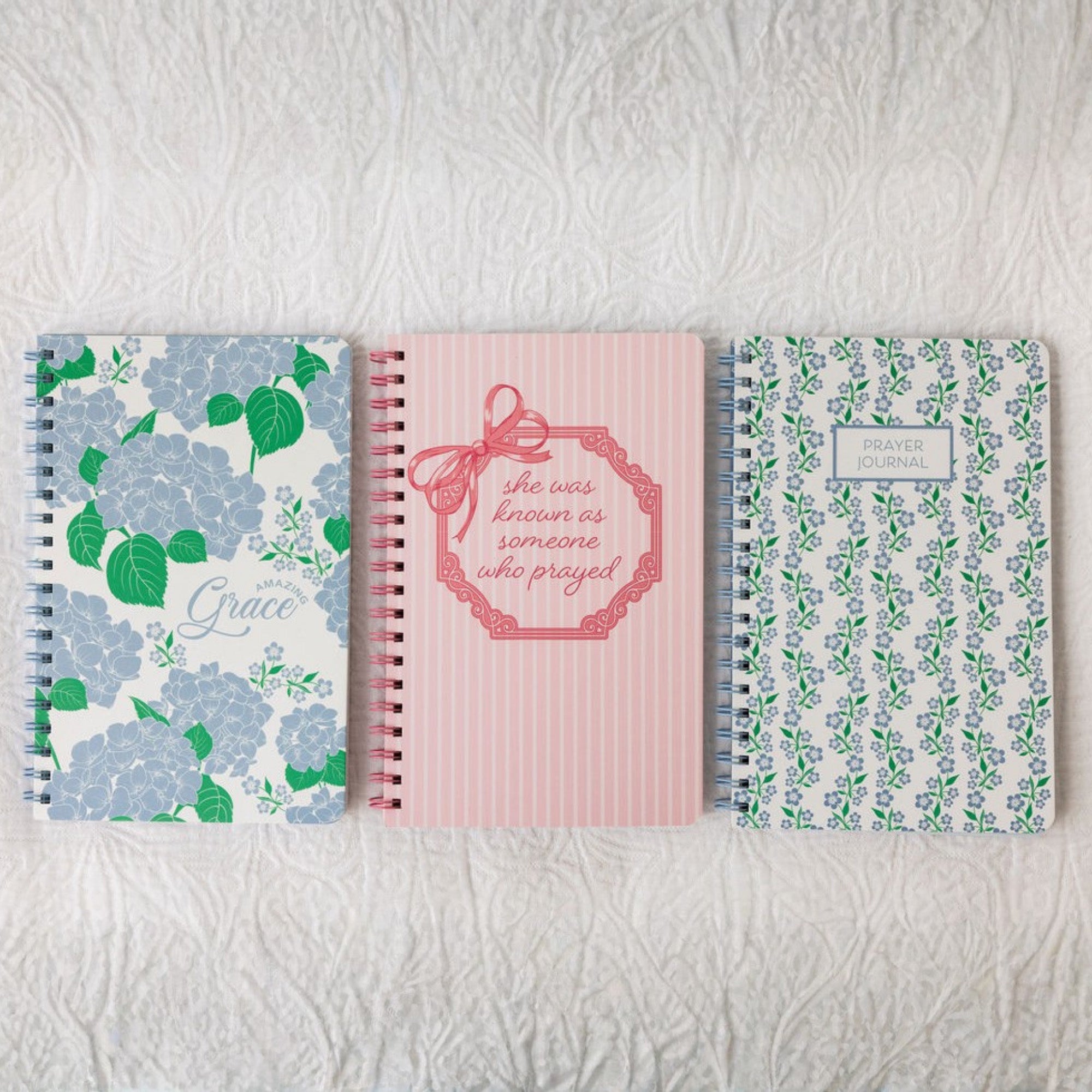 Three spiral-bound notebooks with floral and text designs on a textured white surface.