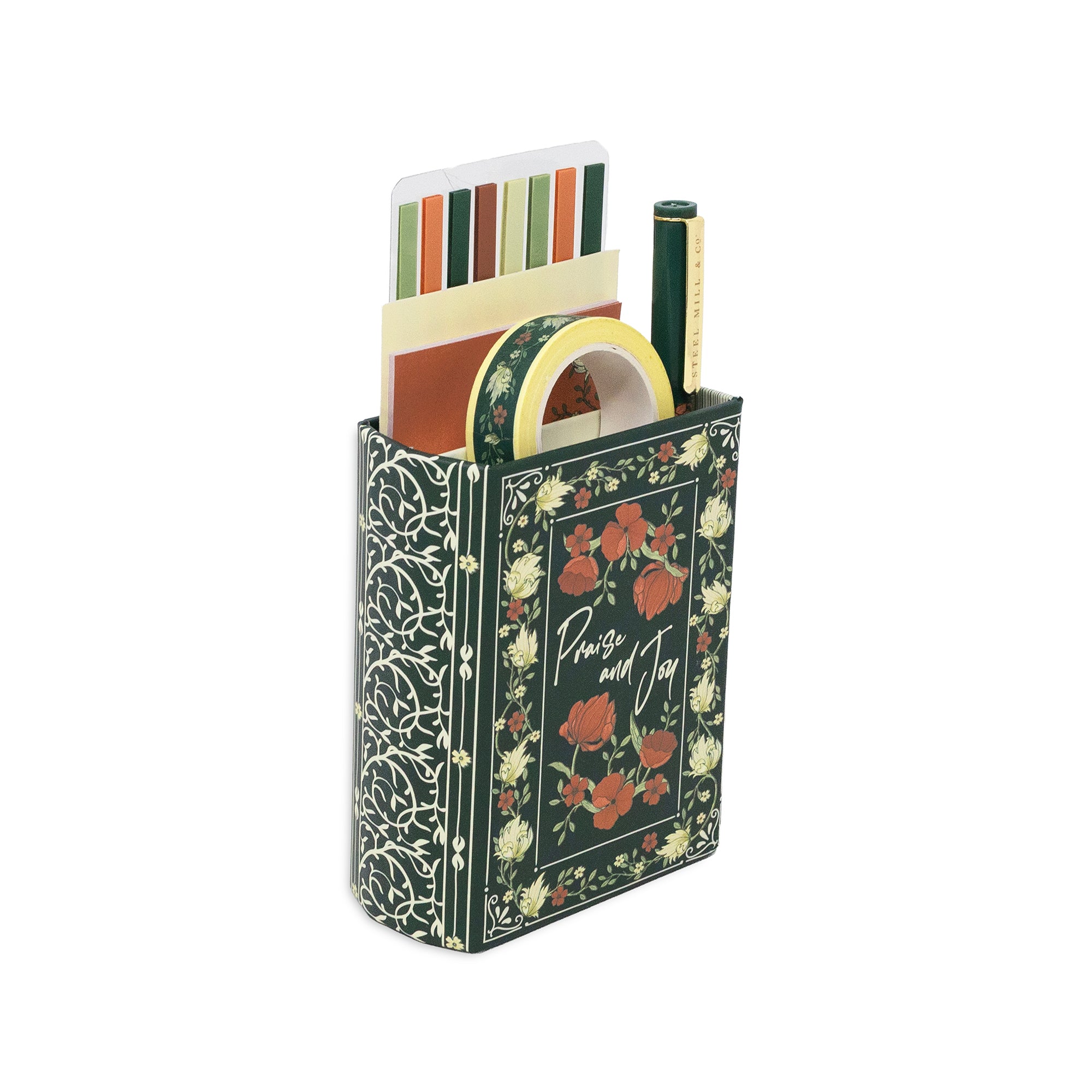 A decorative desk organizer shaped like a vintage book, featuring a dark green cover with red floral illustrations and gold lettering that reads 'Praise and Joy'. The book-style holder is filled with stationery items, including a striped notepad, a roll of washi tape, and pens, all set against a plain white background.