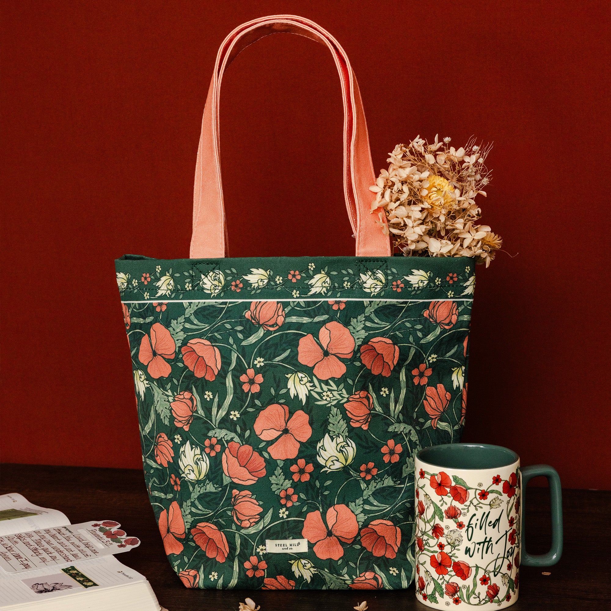 A dark green tote bag with a red and white floral print, featuring coral-pink handles, styled on a dark wooden surface against a deep red background with a ceramic mug and dried flowers as props.