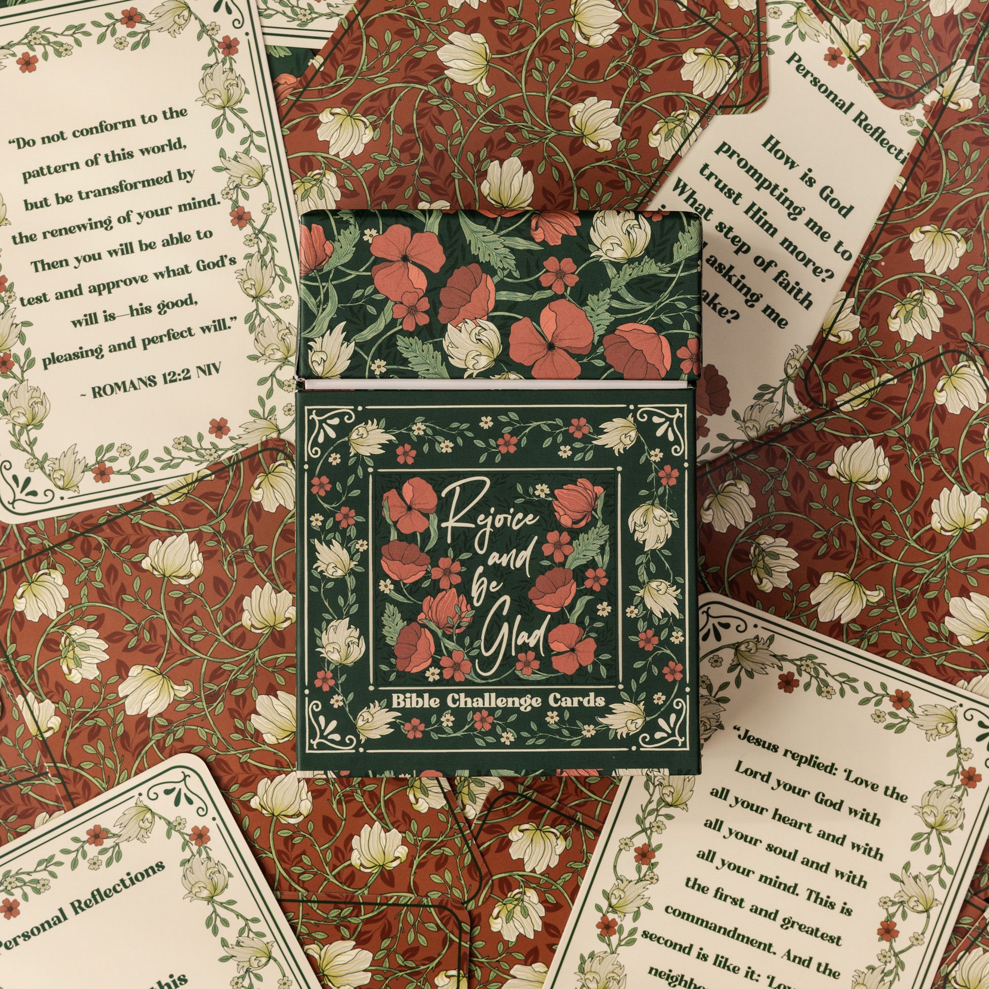 A set of Bible Challenge Cards arranged in a styled flat-lay. The central product is a dark green box decorated with red flowers and green leaves, surrounded by matching patterned cards showing inspirational Bible verses.
