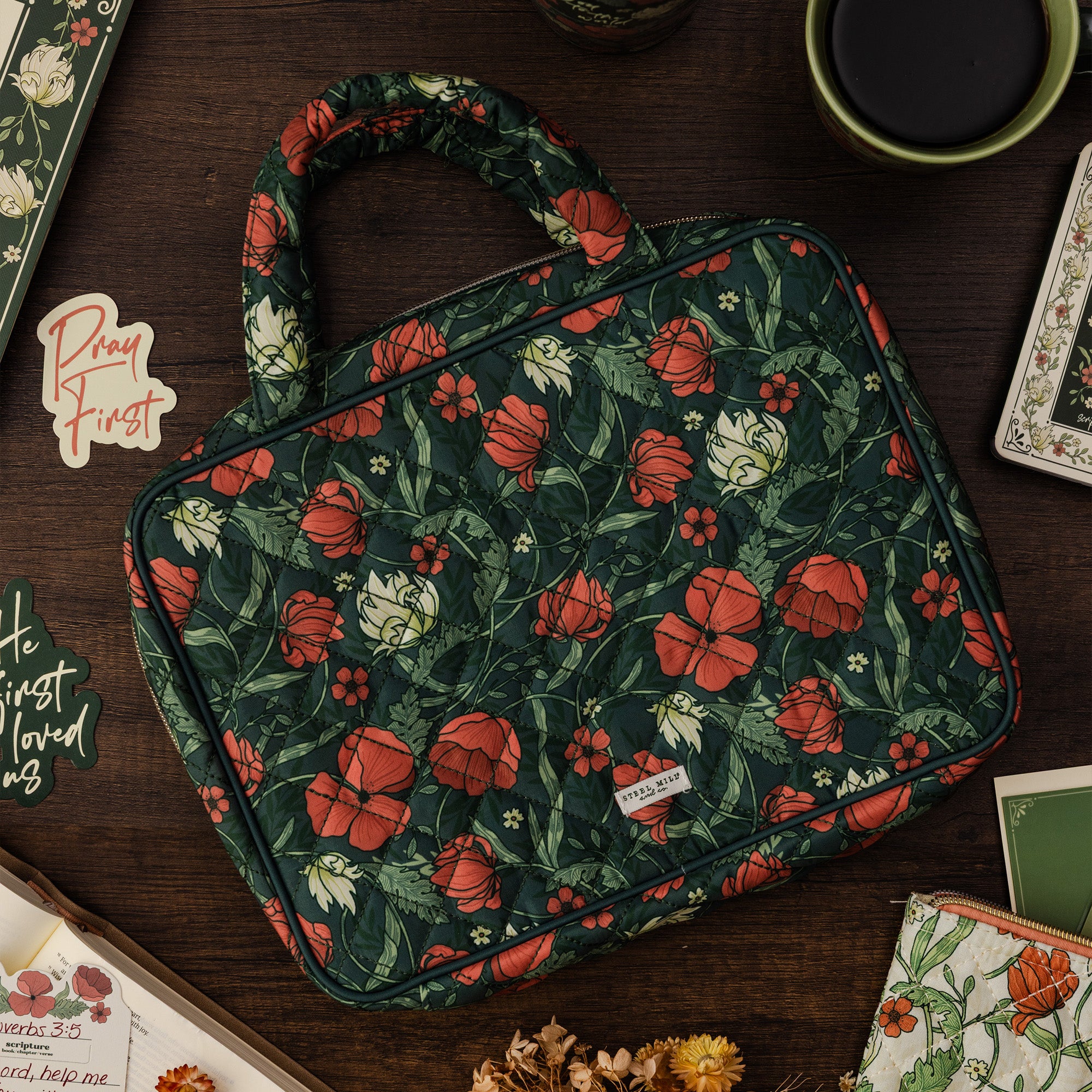 A structured tote-style bag with a dark green floral print, featuring red and white flowers, resting on a dark wooden desk surrounded by styled props.