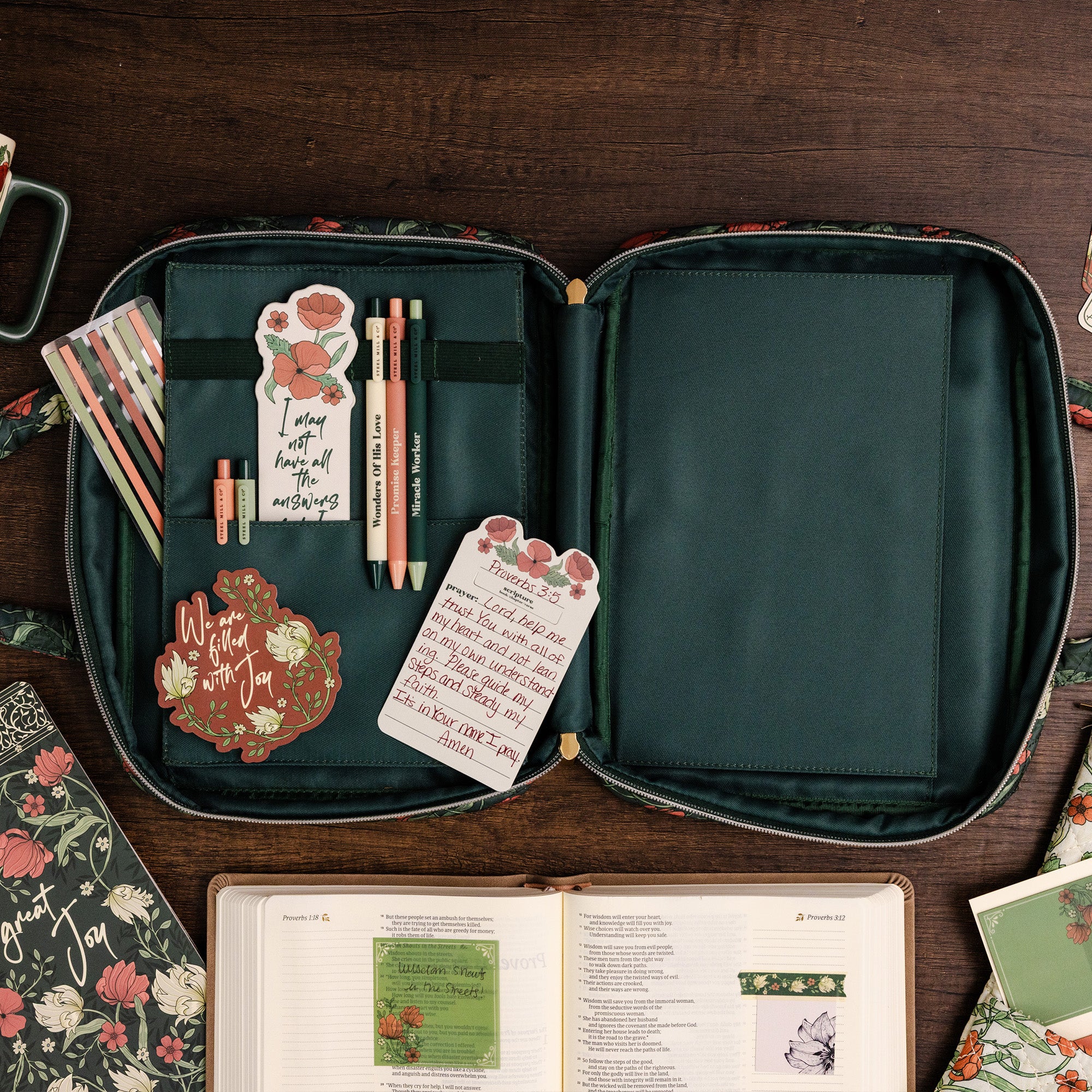An open floral fabric organizer case laid flat on a dark wood surface, holding pens, sticky notes, and small accessories. Surrounding props include a notebook, a book, and other stationery items, creating a styled flat-lay scene.