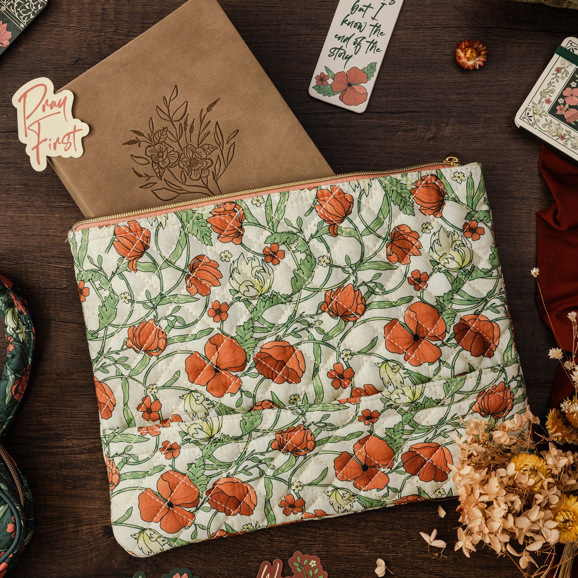 A quilted fabric pouch with an orange poppy and green vine floral pattern, styled on a dark wood background with a kraft paper notebook, dried flowers, and other cozy flat-lay props.