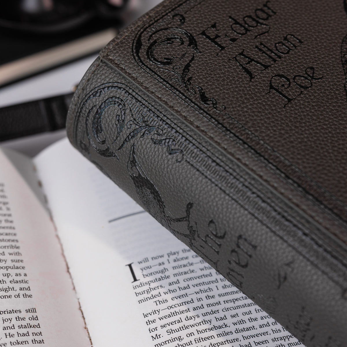 A close-up of a dark brown textured crossbody, showing the embossed title 'The Raven' on the cover, resting on an open book.