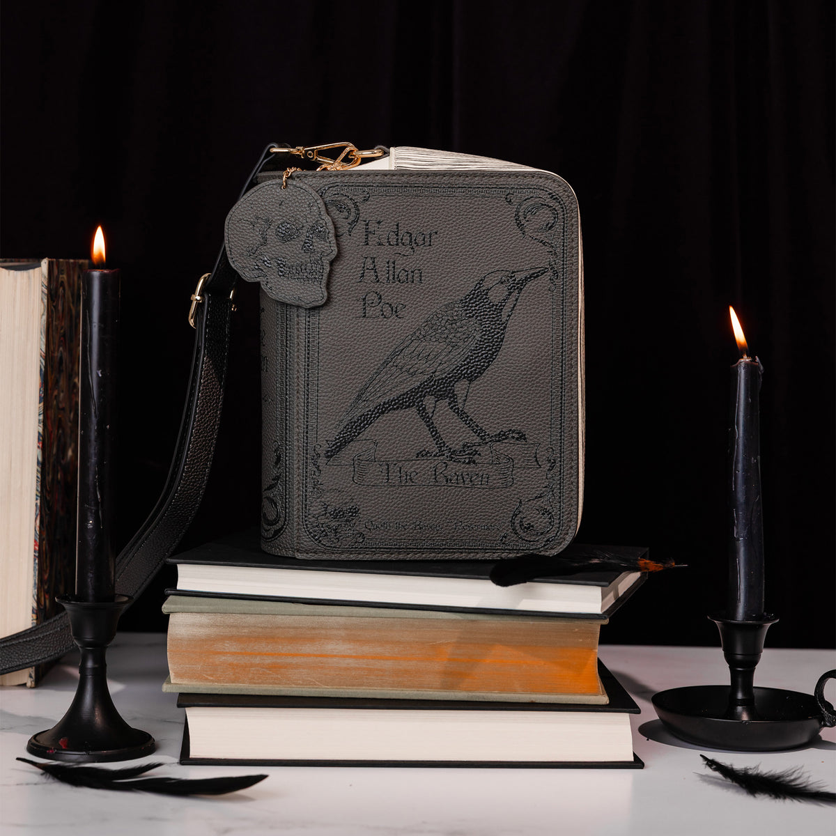 A dark gray faux-leather crossbody bag styled on a stack of books, flanked by lit black candles and a feather, against a black background. The bag features an embossed raven and the text 'Edgar Allan Poe' and 'The Raven'.