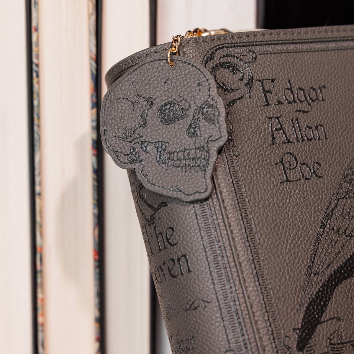 A close-up of a gray faux-leather crossbody featuring a skull-shaped zipper pull, embossed gothic lettering, and a gold zipper pull, set against a background of books.