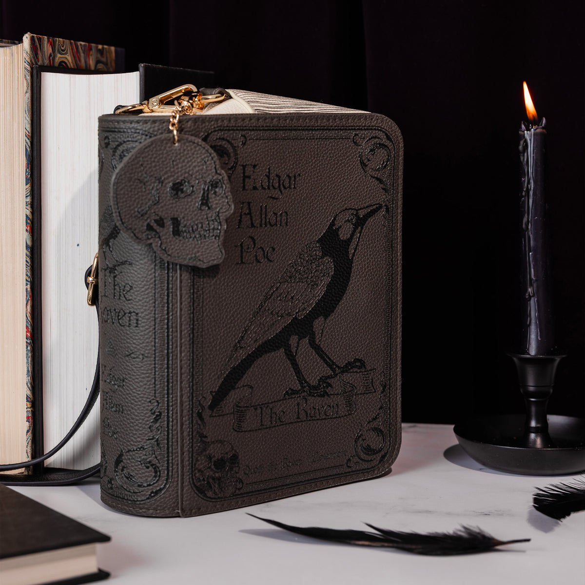 A dark brown faux-leather book-style clutch or organizer with 'The Raven' Edgar Allan Poe design, styled on a tabletop with books, a lit candle, and feathers against a black background.
