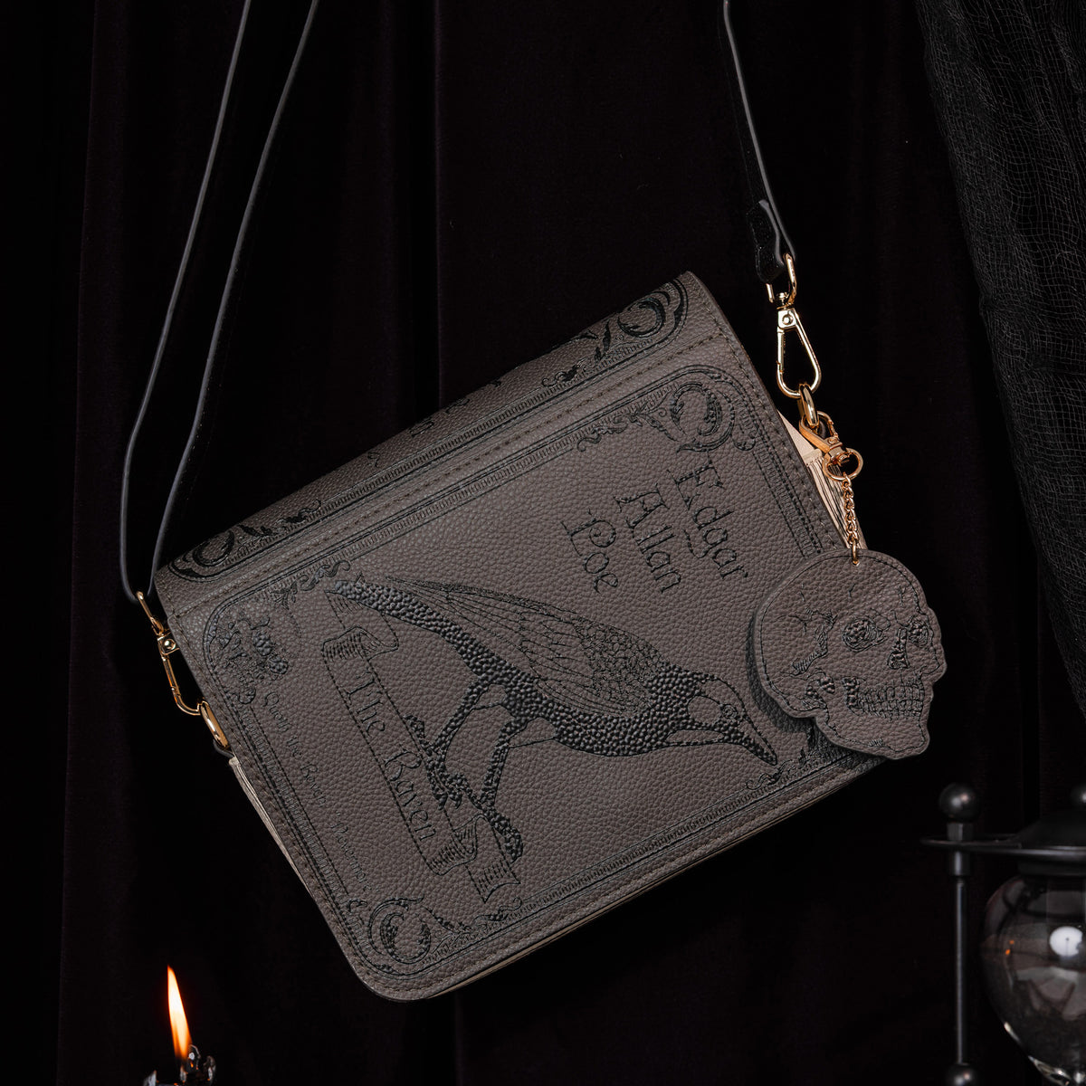 A dark gray faux-leather crossbody bag styled like an old book cover, featuring embossed gothic text, a raven illustration, and gold-tone hardware, shown against a dark draped background.
