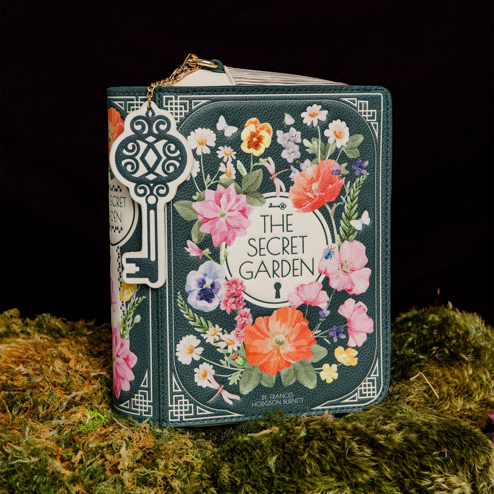 A decorative book-shaped bag styled like 'The Secret Garden' book cover, featuring a floral wreath illustration and a large ornate key charm, displayed on moss against a black background.