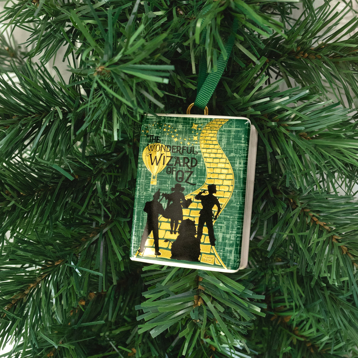 A small decorative ornament shaped like the book 'The Wonderful Wizard of Oz.' with a green ribbon loop at the top, shown on a holiday tree