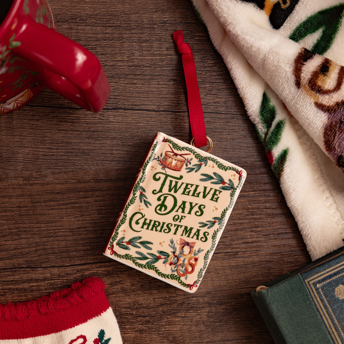 A Christmas book ornament featuring the title 'Twelve Days of Christmas' in decorative lettering, with a red hanging ribbon and a small gold loop at the top, shown on a table