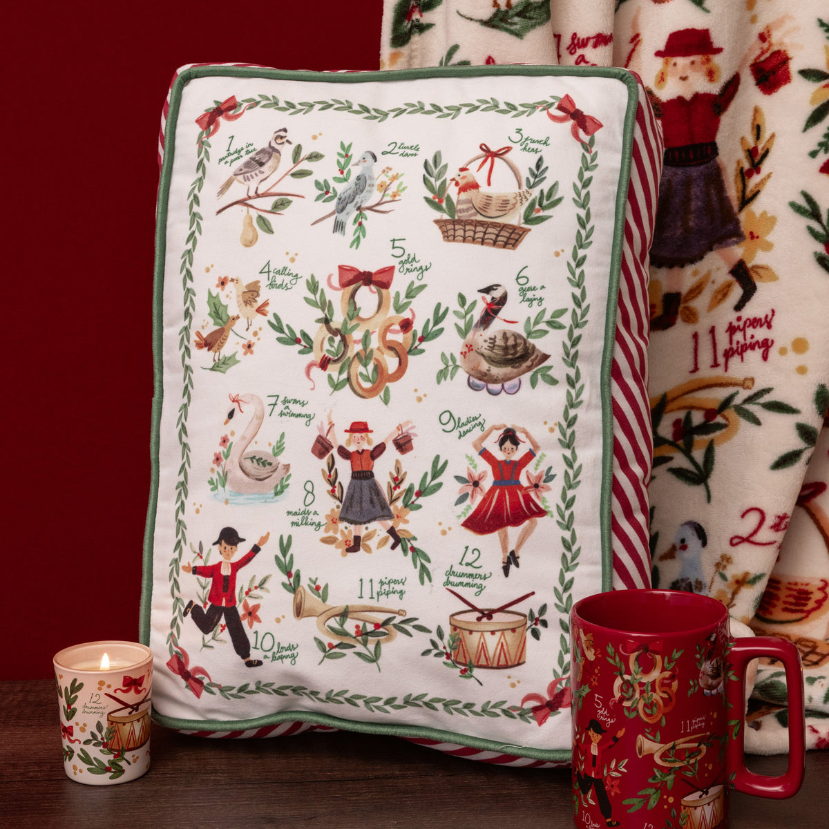 A rectangular decorative pillow with a Christmas-themed illustrated calendar design, styled on a dark wooden surface next to a lit candle, a red mug, and a matching patterned blanket against a dark red background.
