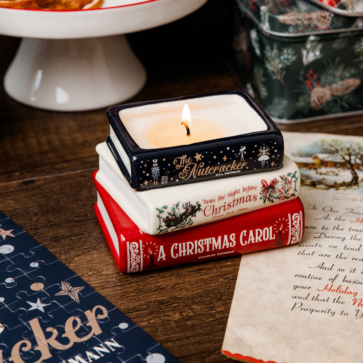 A Christmas-themed candle holder designed to look like a stack of three book spines reading 'The Nutcracker', 'twas the night before Christmas', and 'A Christmas Carol', with a lit white candle on top, styled on a wooden tabletop with holiday props.