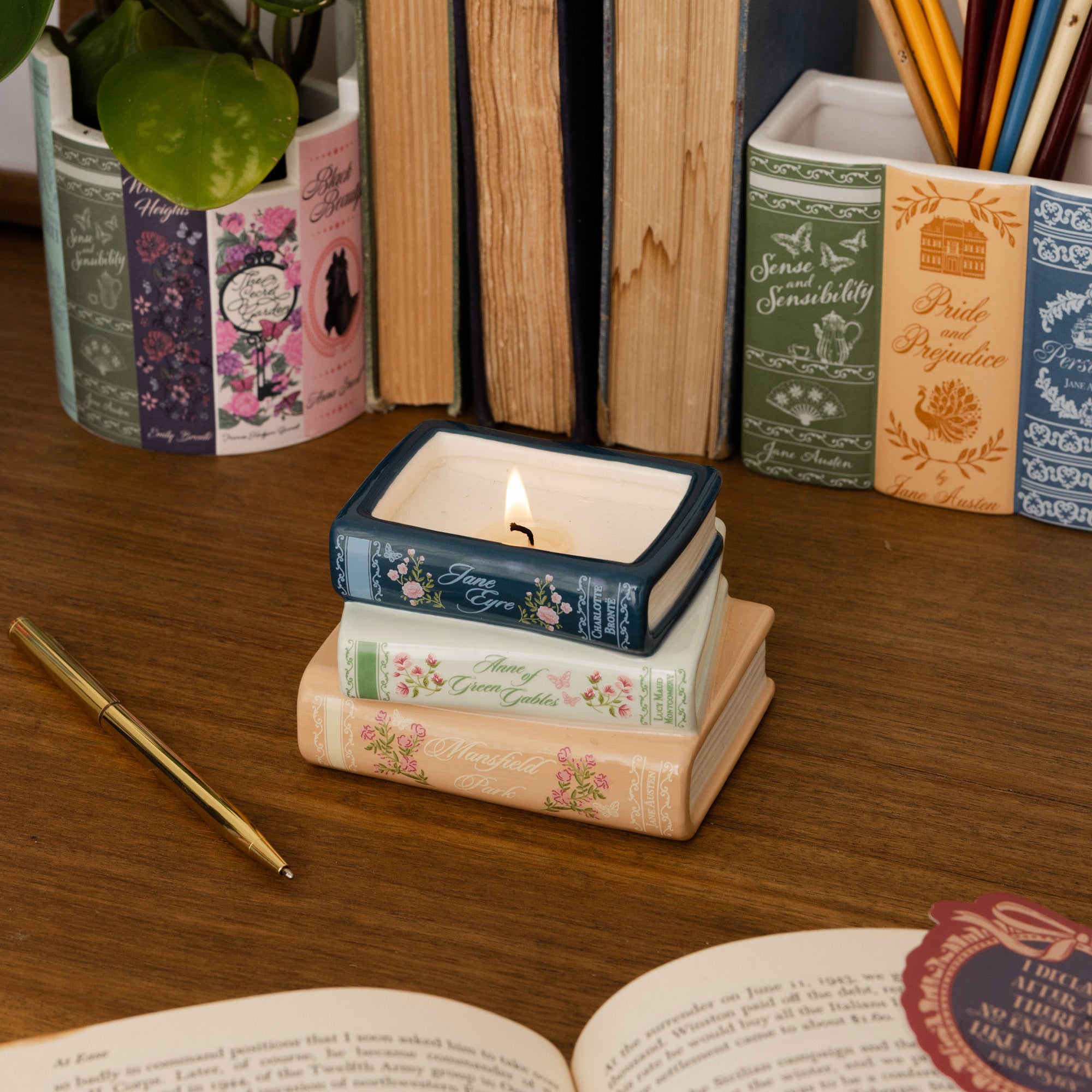 A stack of three book-shaped candles sits on a wooden desk, styled with books, a pen, pencils, and a plant. The candles are designed to look like vintage books with floral patterns and are lit.