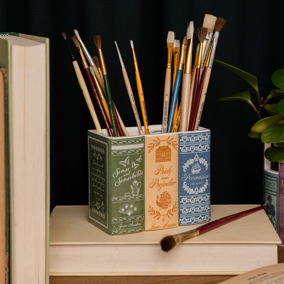 A rectangular ceramic planter or container with a three-spine book design. The left spine is dark green with white script and botanical illustrations, the middle spine is tan with gold script and a house illustration, and the right spine is blue with white decorative patterns.