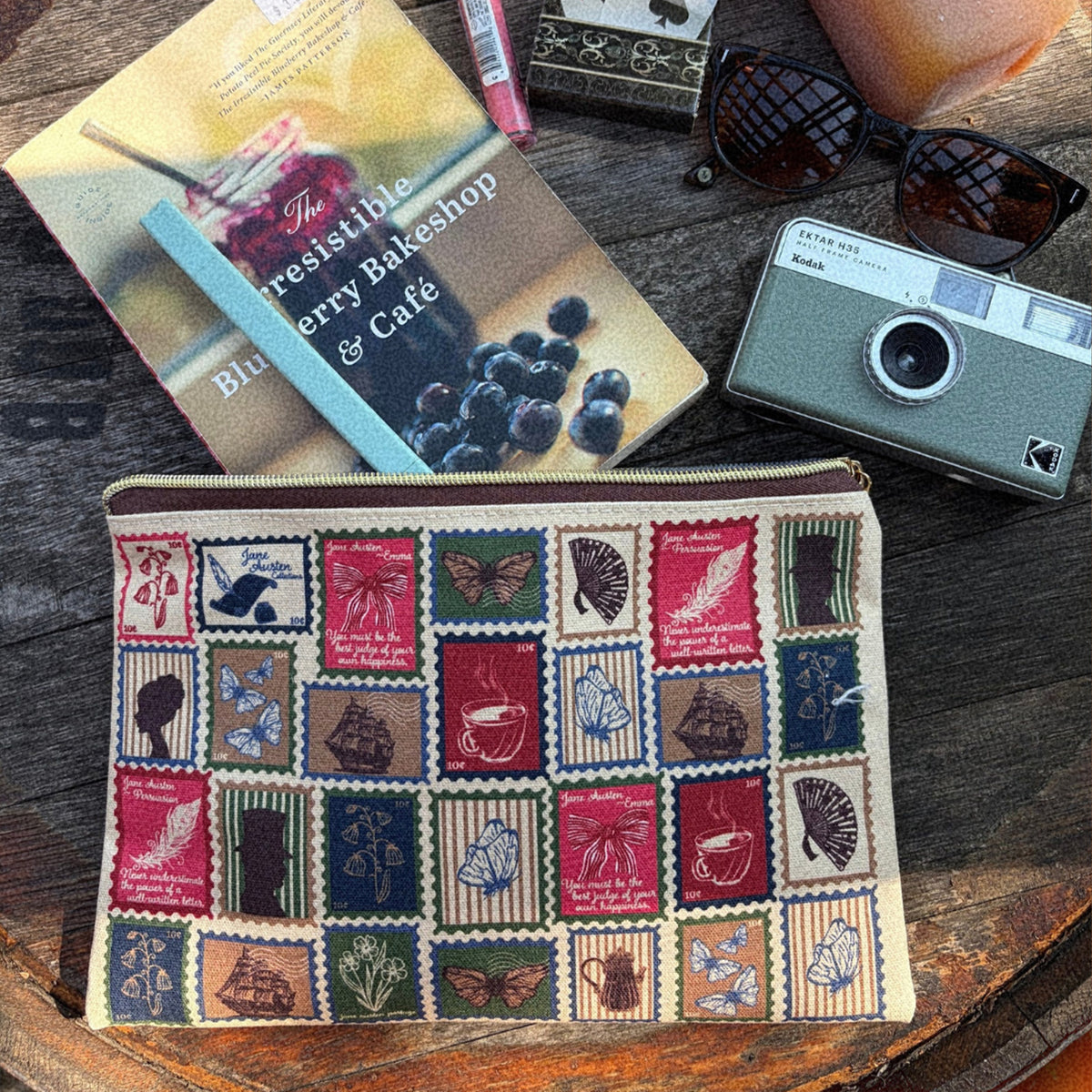A fabric zippered pouch with a vintage postage stamp patchwork pattern, styled on a rustic wooden table alongside a book, sunglasses, a camera, and sunglasses