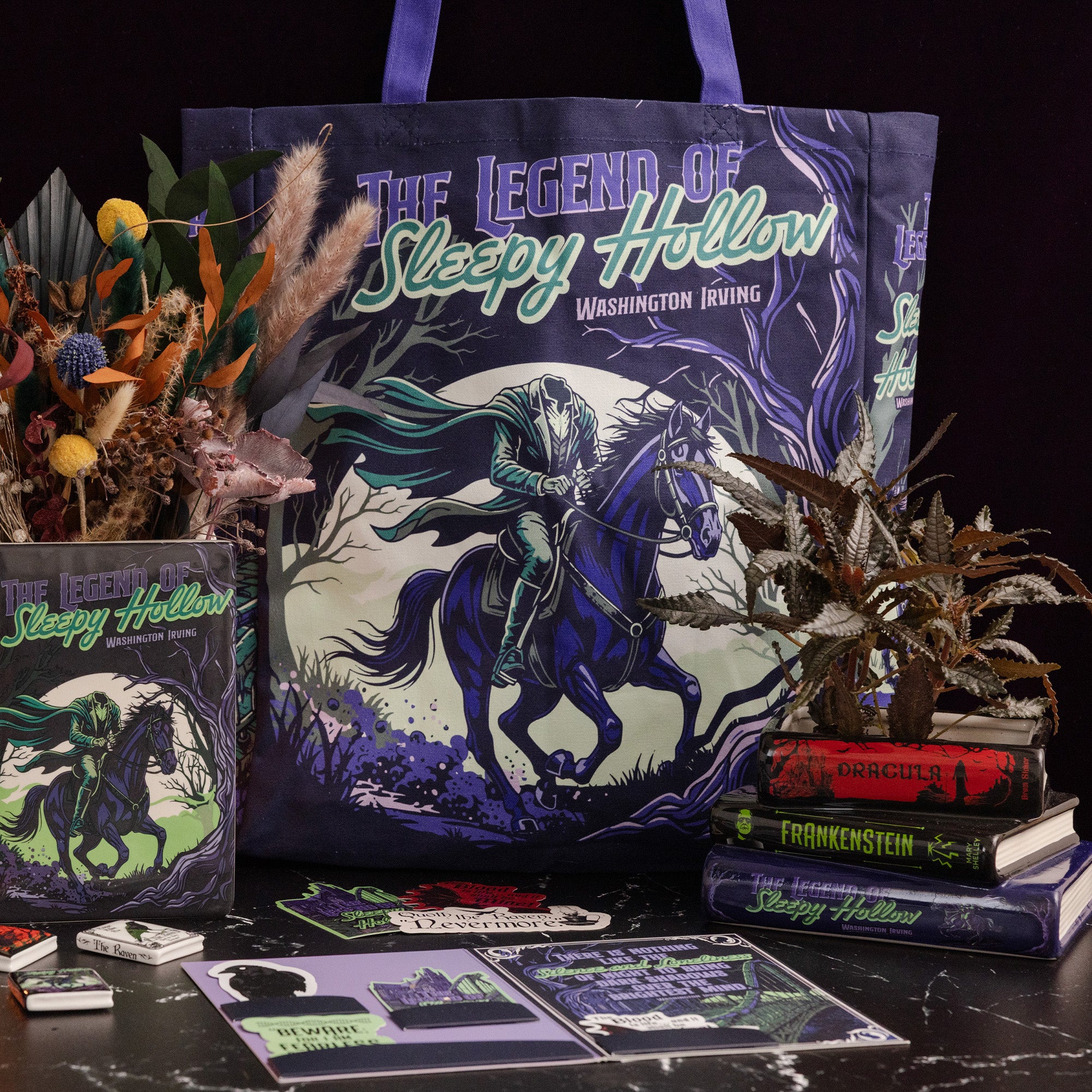 A themed tote bag and matching printed cards, stickers, and book-style items from The Legend of Sleepy Hollow, arranged on a dark tabletop with decorative dried flowers and books against a black background.
