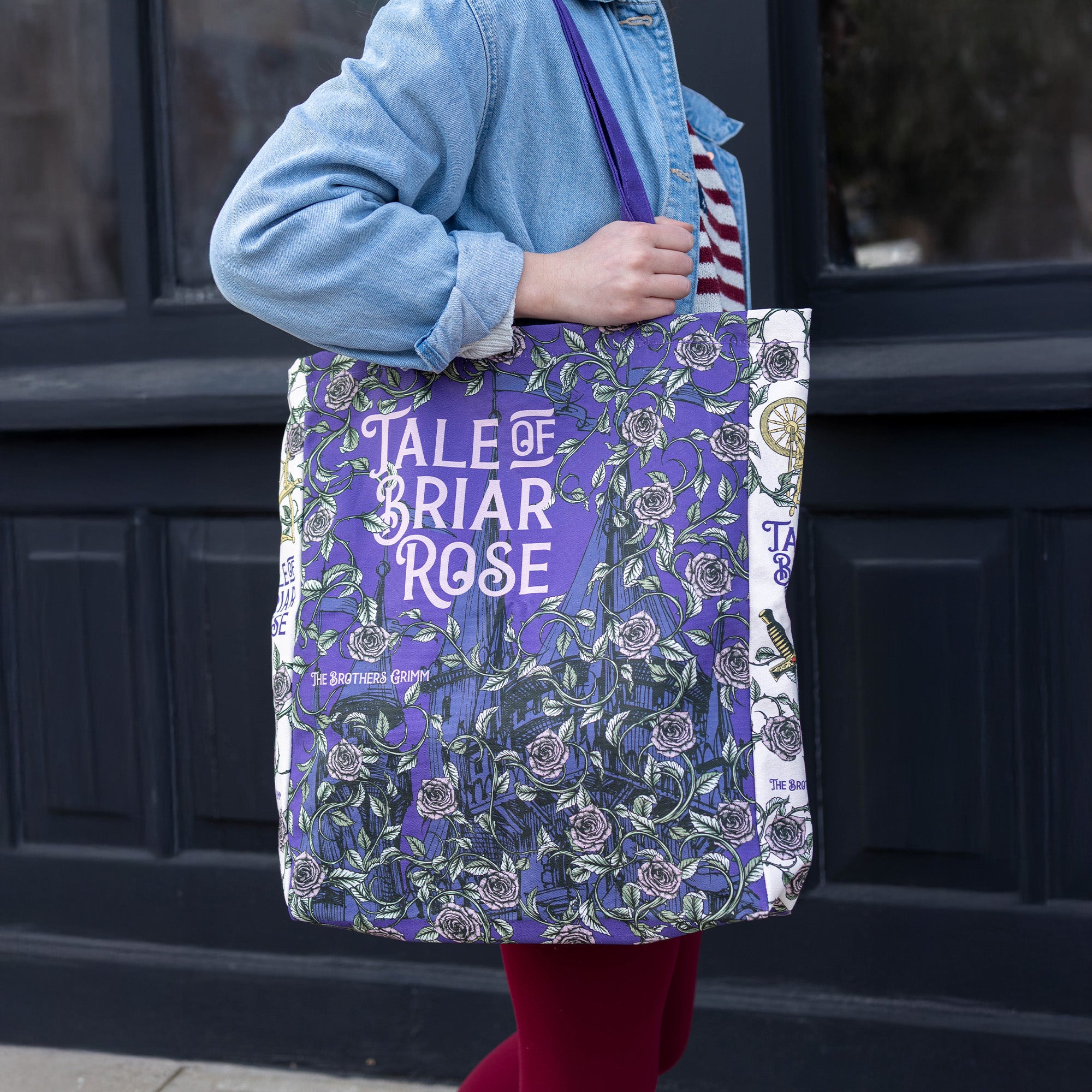 Person holding a purple tote bag with floral design and text in front of a dark door.