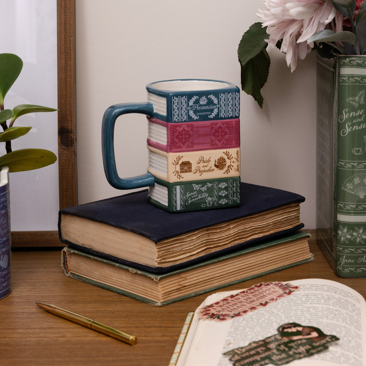 A ceramic mug designed to look like a stack of vintage books, featuring a teal handle and multicolored book-spine patterns, displayed on a stack of old books on a wooden desk.
