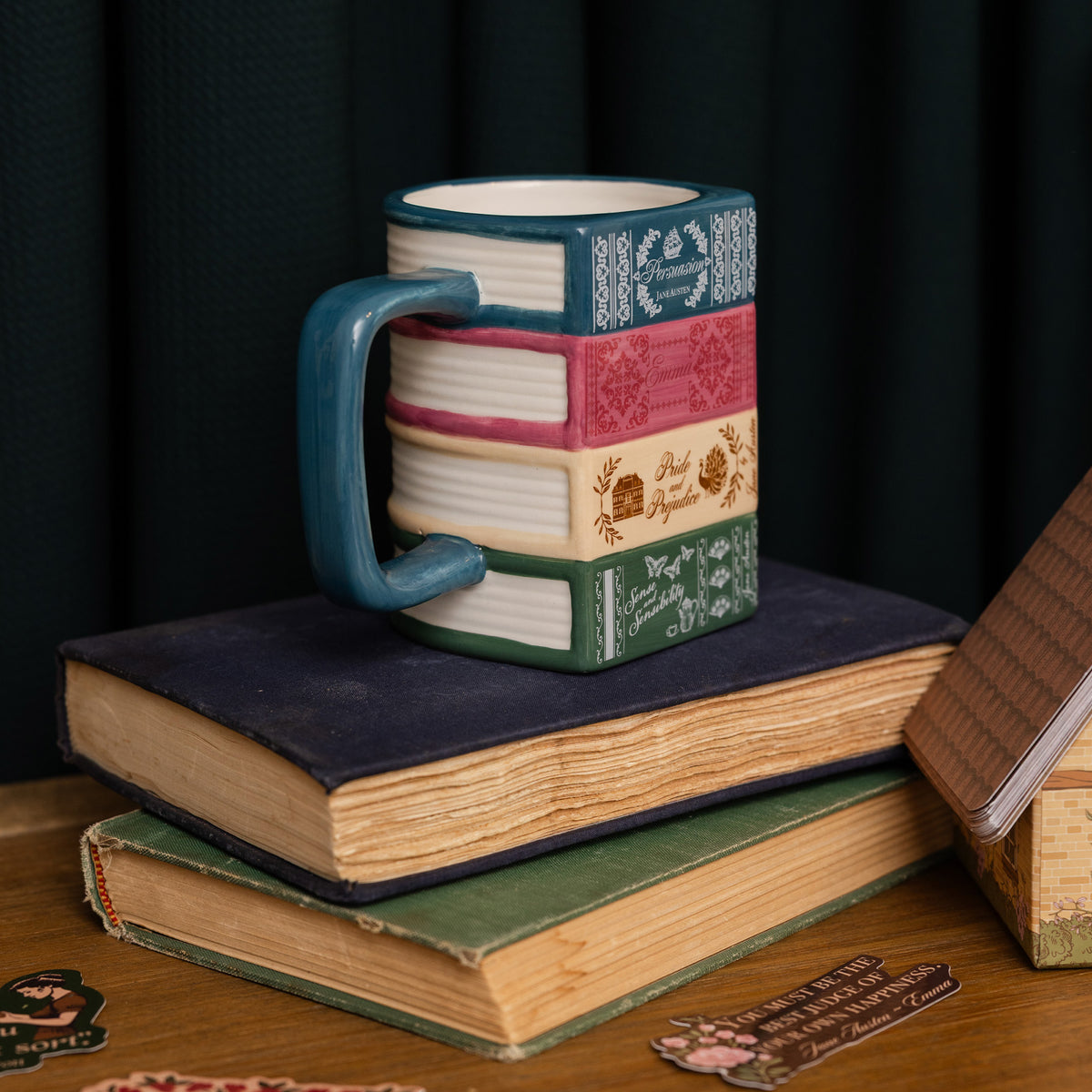 A ceramic mug shaped like a stack of vintage books with a teal handle, displayed on top of stacked antique books against a dark curtain background.