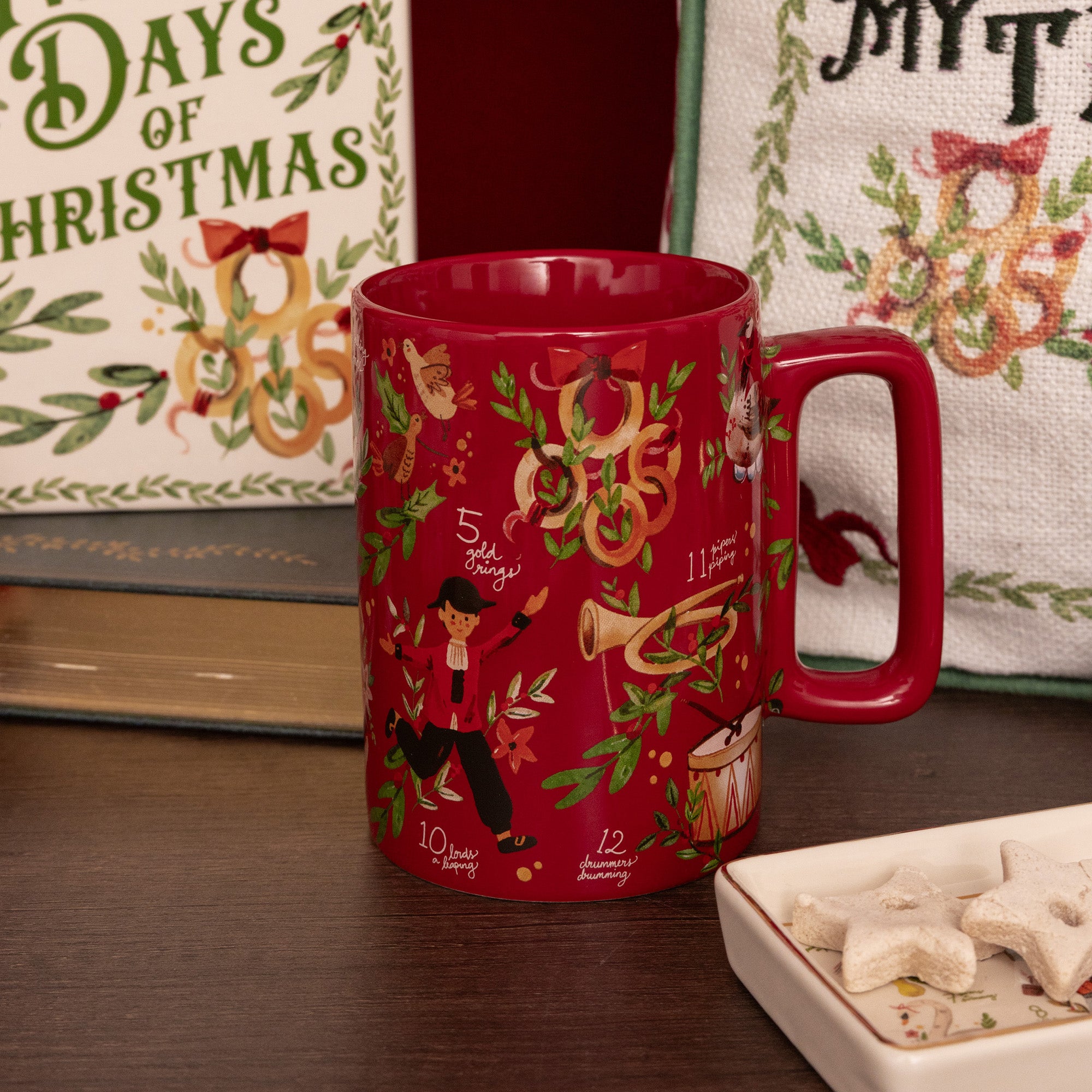 A red ceramic mug with a holiday music theme, featuring illustrations of musical instruments, notes, and a dancer, displayed on a wooden table with Christmas-themed background decor.