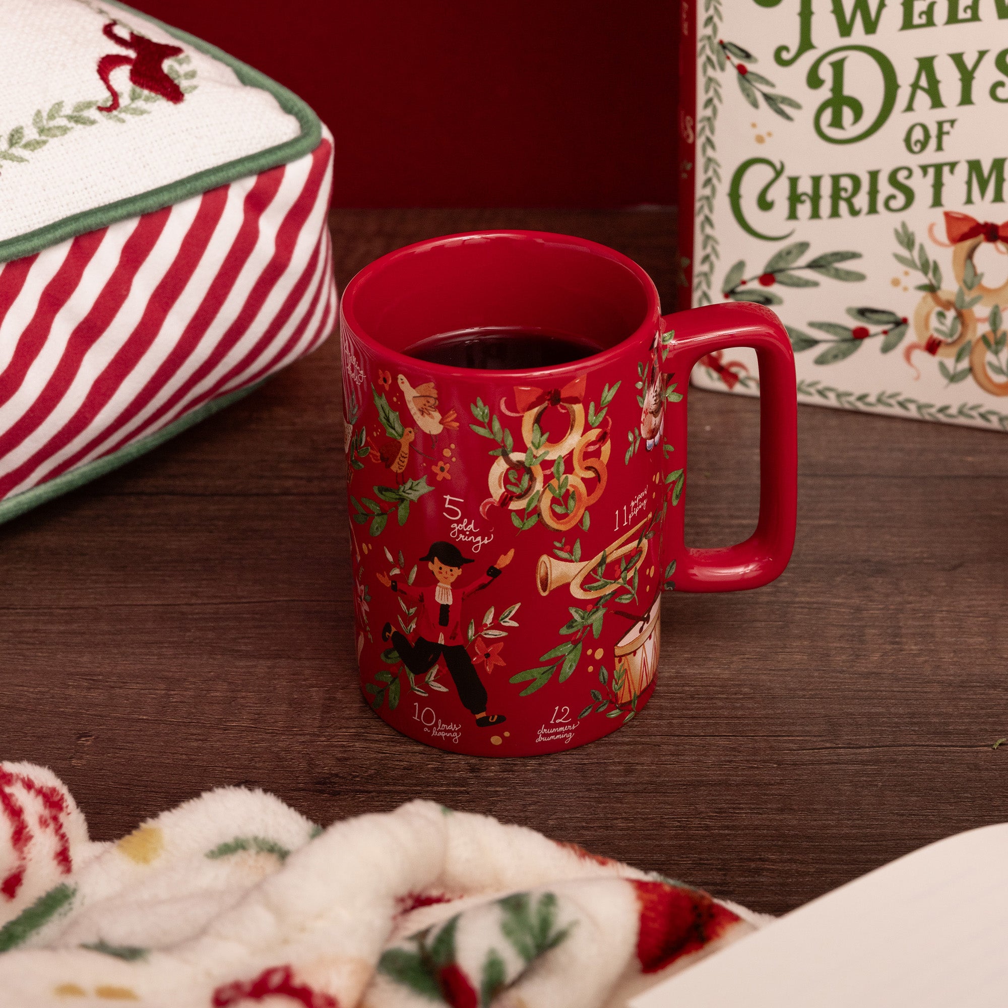 A festive red ceramic mug with a holiday illustration of Santa Claus and Christmas ornaments, sitting on a dark wooden table surrounded by cozy Christmas-themed props.