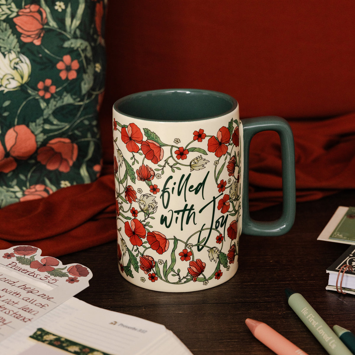 A ceramic mug with a cream-colored exterior decorated with red poppies and green leaves, featuring the cursive text 'Filled with Joy' in black. The mug has a dark teal interior and handle, and is styled on a dark wooden table with a floral pillow, red fabric, and stationery in the background.
