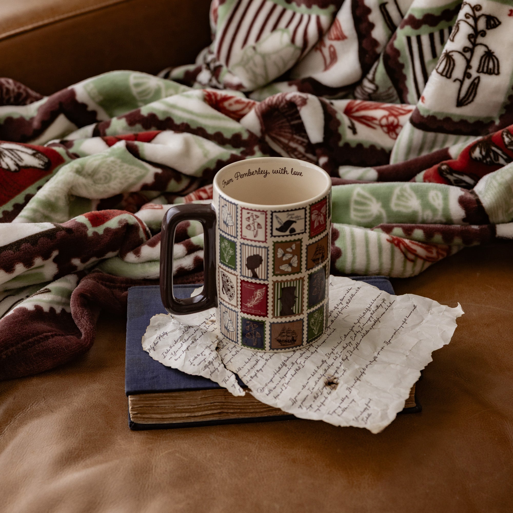 A ceramic mug with a dark handle sits on a vintage book and torn paper, surrounded by a cozy patterned blanket on a brown leather couch. The mug features a patchwork design of small illustrated squares with the text 'Jim Frederickly with love' printed inside.