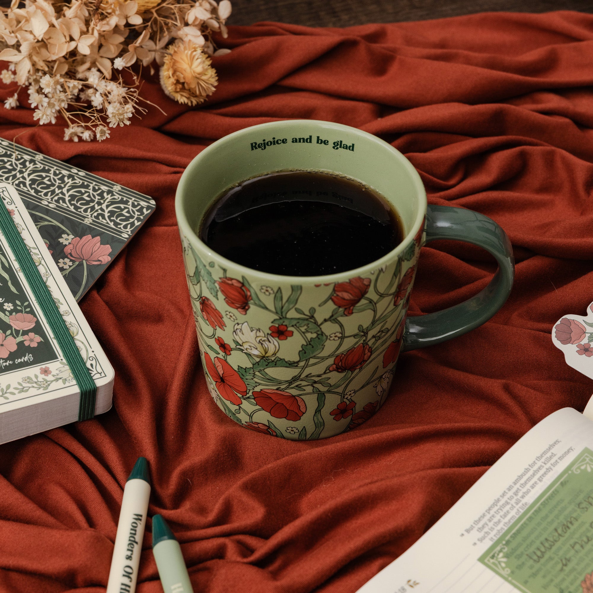 A ceramic mug filled with black coffee sits on rust-colored fabric, surrounded by dried flowers, notebooks, pens, and an open book. The mug has a pale green base with a red floral pattern and the words 'Rejoice and be glad' printed inside near the rim.