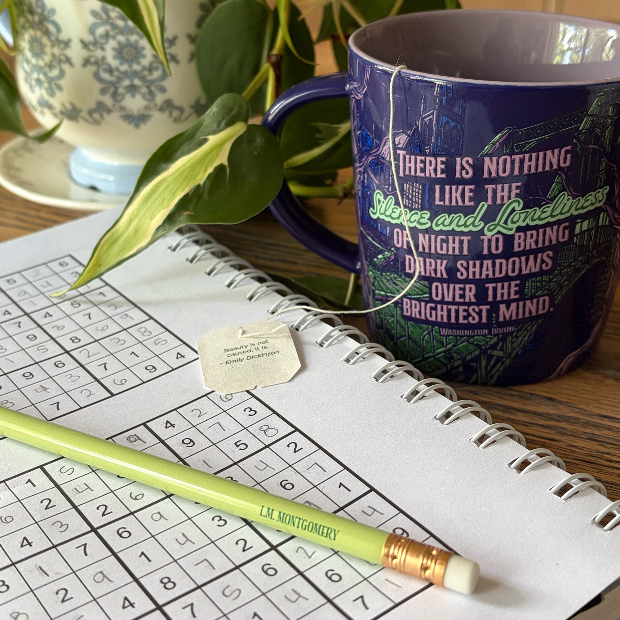 A dark blue ceramic mug with a quote printed on it sits on a wooden table next to an open spiral notebook with a crossword puzzle, a green pencil, and a plant leaf in the foreground.