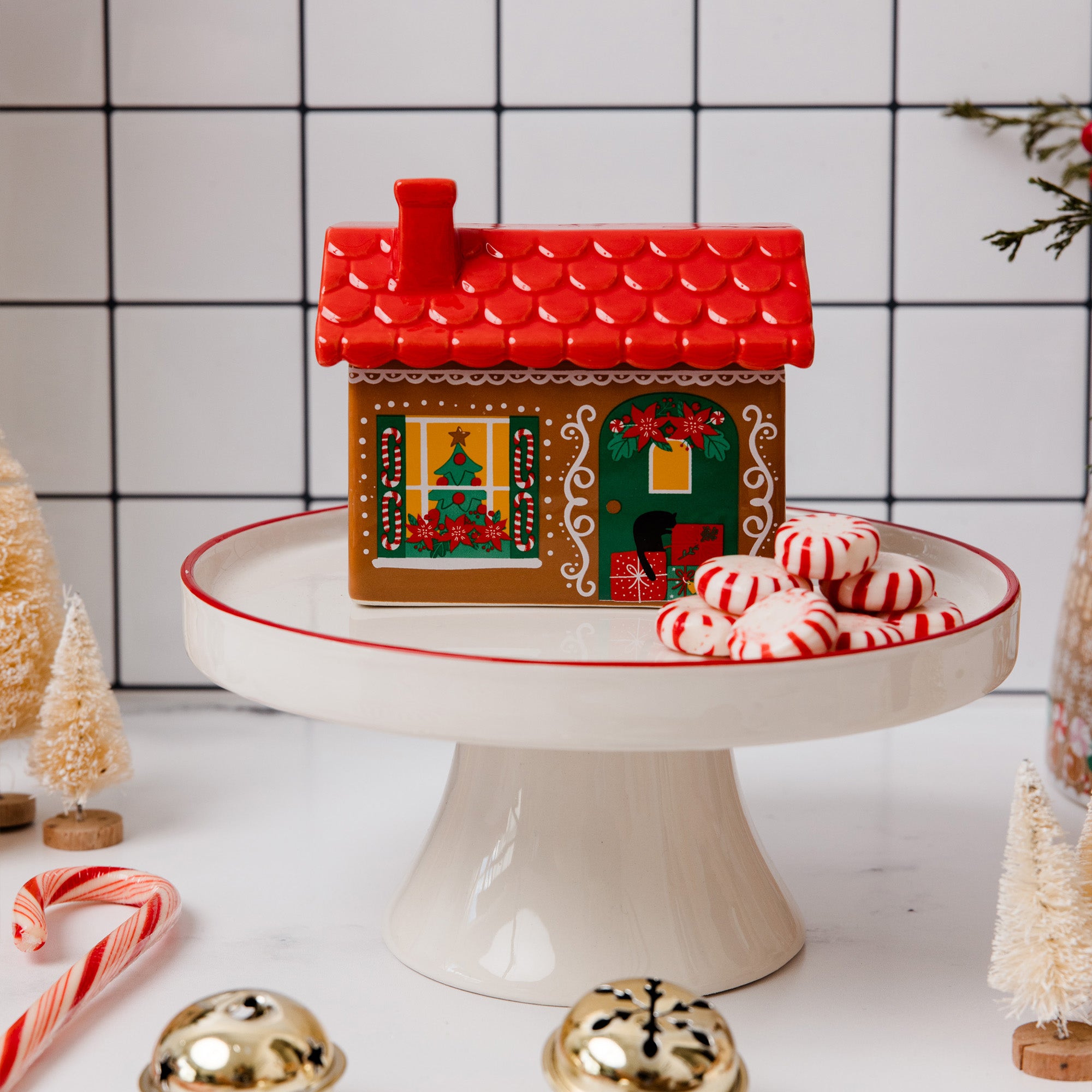 A ceramic gingerbread decorative house sits on a white pedestal cake stand, surrounded by candy canes, gold bells, and miniature Christmas trees against a white tiled background.