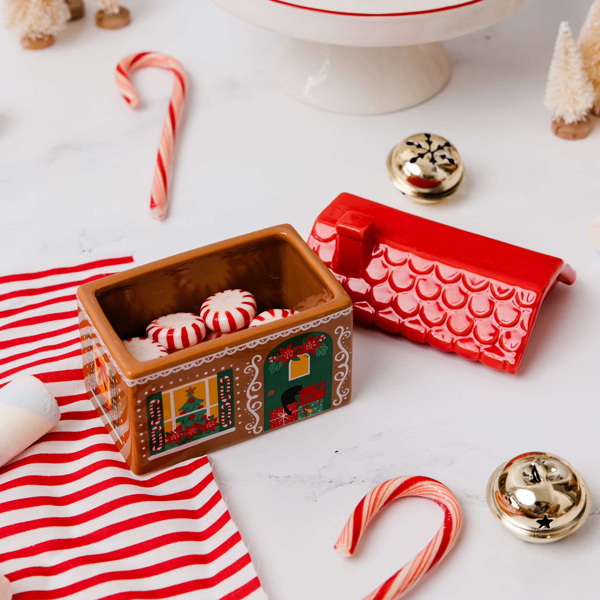 A Christmas-themed ceramic candy box set styled on a white marble surface with candy canes, gold bells, and a striped napkin. The set includes a gingerbread house-shaped box filled with peppermint candies, a red house-shaped box, and two gold bells.