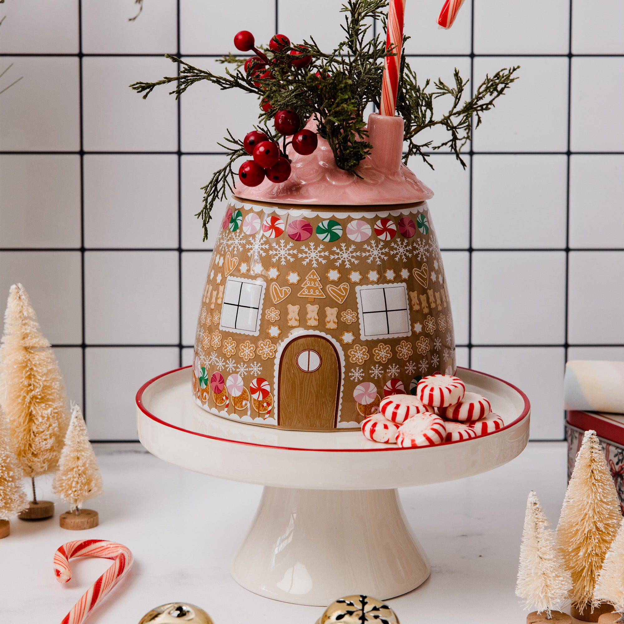 A gingerbread house-shaped ceramic jar or container with a pink lid, decorated with white snowflakes, candy canes, and a brown door, displayed on a white pedestal stand with peppermint candies and small decorative trees.