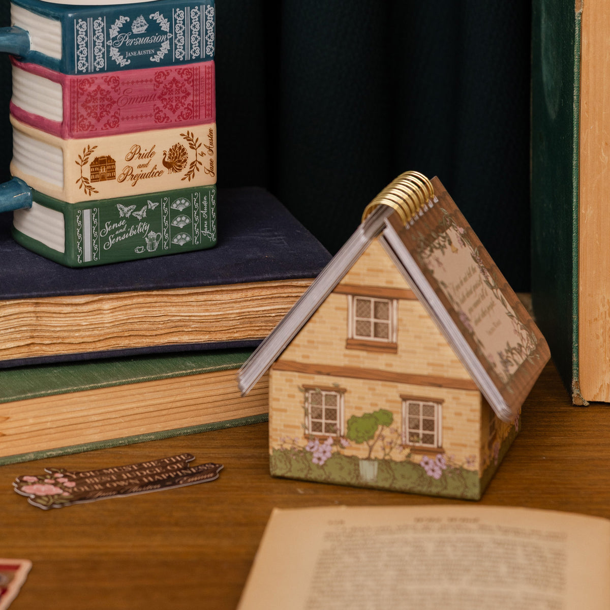 A miniature house-shaped inspirational quotes with a spiral top sits on a wooden surface next to stacked vintage books. The house features a beige brick pattern, green foliage, purple flowers, and a gold spiral binding.