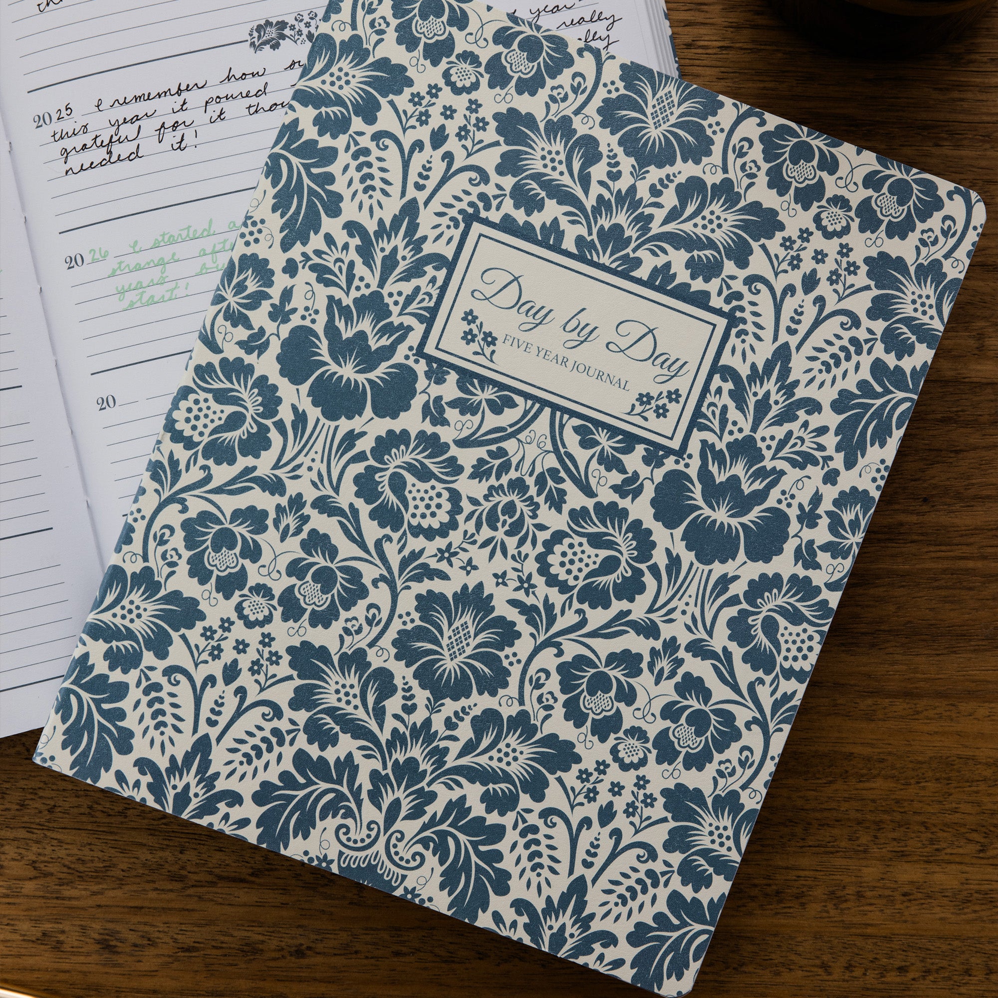 A floral patterned five-year journal titled 'Day by Day' lies open on a wooden surface, with handwritten notes visible on the left page.