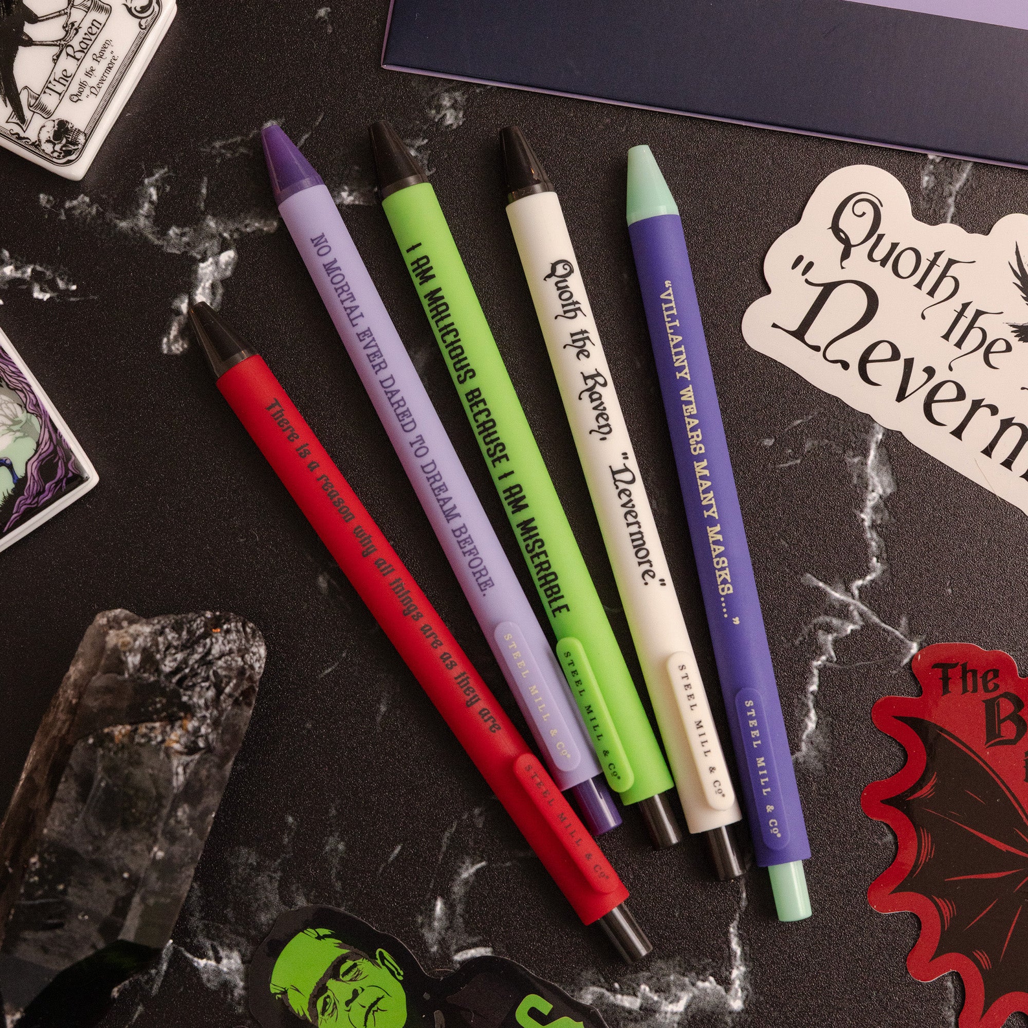 Five pens in assorted colors (red, purple, green, cream, and dark blue) with gothic-themed text, arranged on a dark marble surface