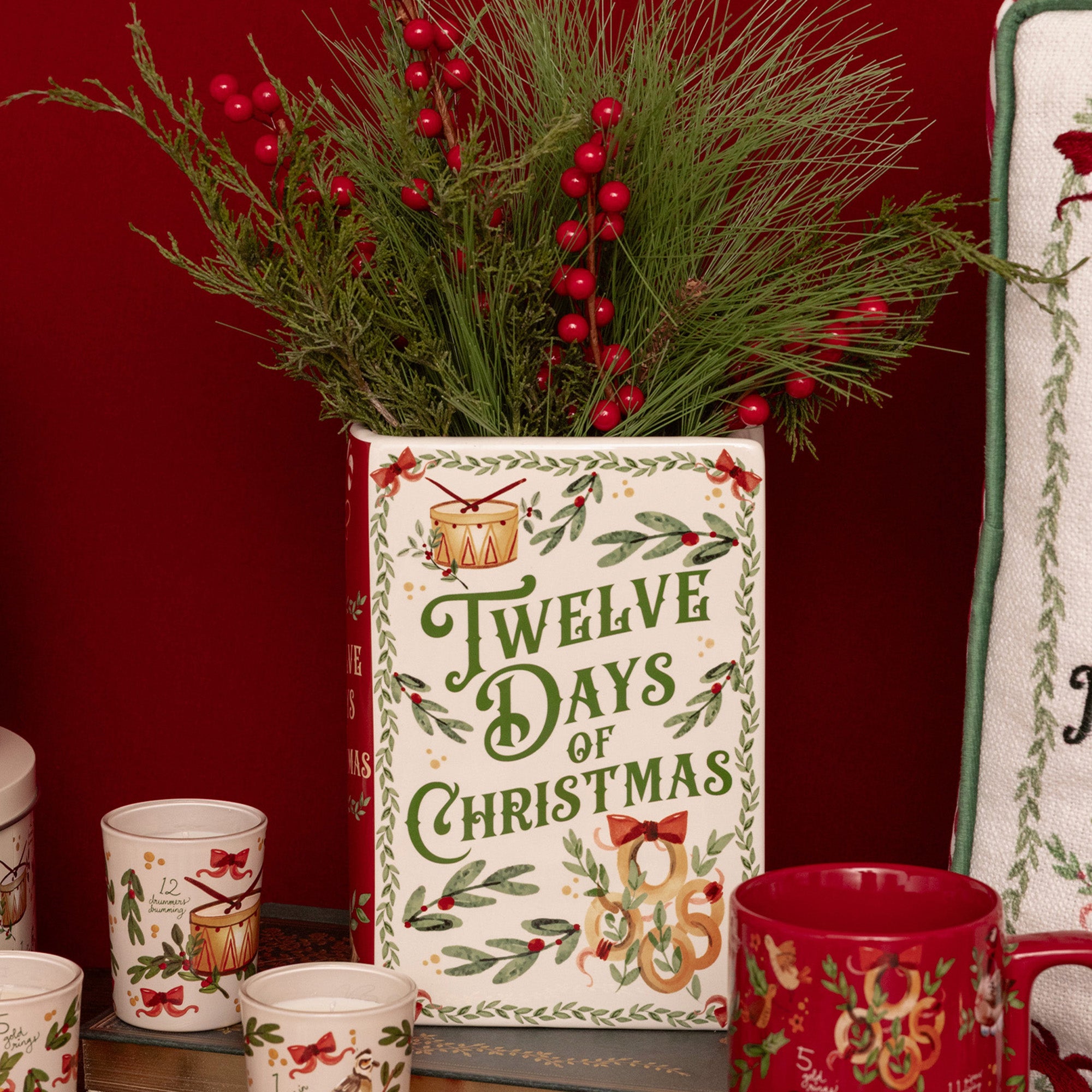 A festive Christmas-themed ceramic planter with 'Twelve Days of Christmas' printed on the front, filled with green pine branches and red berries, styled on a shelf with matching red and white mugs against a dark red background.
