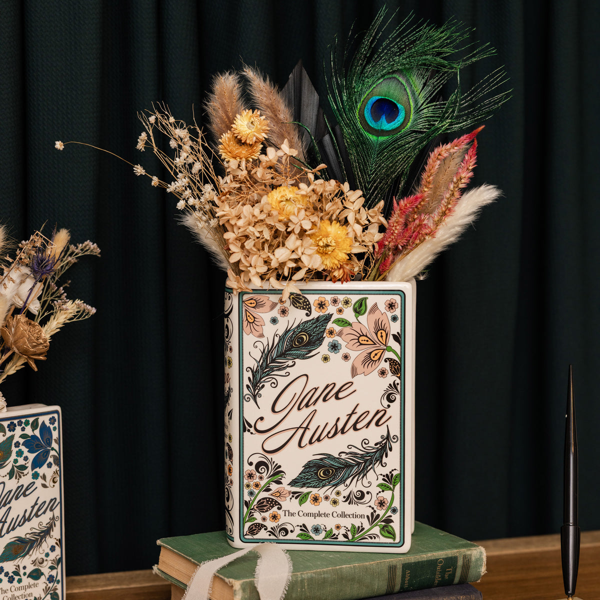 A decorative ceramic vase printed with Jane Austen book covers, filled with dried flowers, pampas grass, and a peacock feather, styled on books against a dark curtain background.