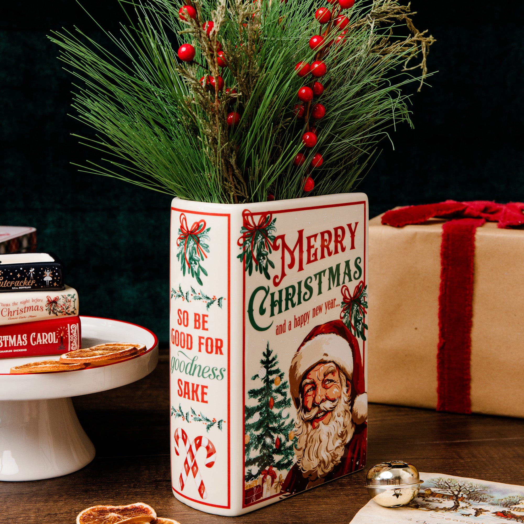 A ceramic Christmas tin canister vase with a festive red and green holiday print, filled with pine branches and red berries, set on a dark wooden table with wrapped gifts in the background.