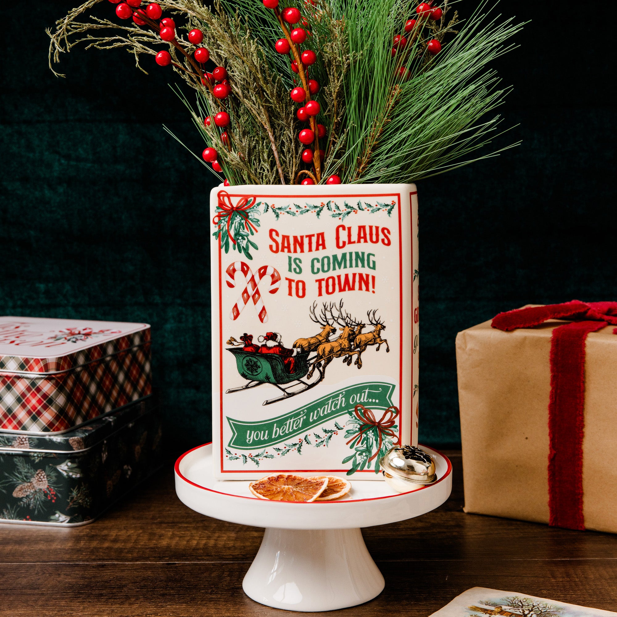 A rectangular ceramic Christmas planter with a festive red and green design reading 'Santa Claus is coming to town!' and 'you better watch out!', displayed on a white pedestal stand with pine branches and red berries placed inside it. Gift boxes and stacked tins sit in the background against a dark backdrop.