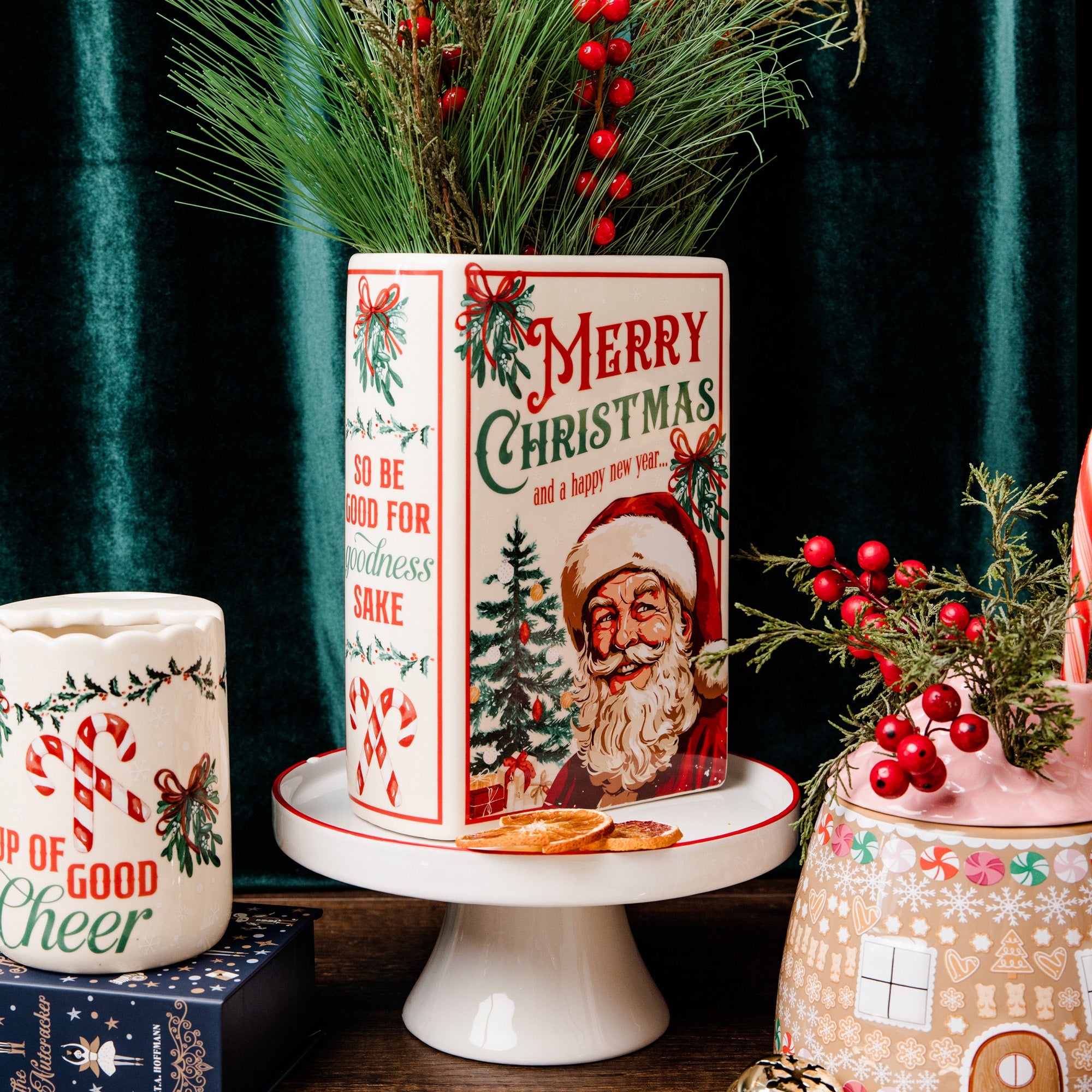 A vintage-style ceramic Christmas vase with a Santa Claus illustration and 'Merry Christmas' text, displayed on a white pedestal stand. The canister is decorated with red berries, pine branches, and holiday greenery, set against a dark green velvet curtain background.