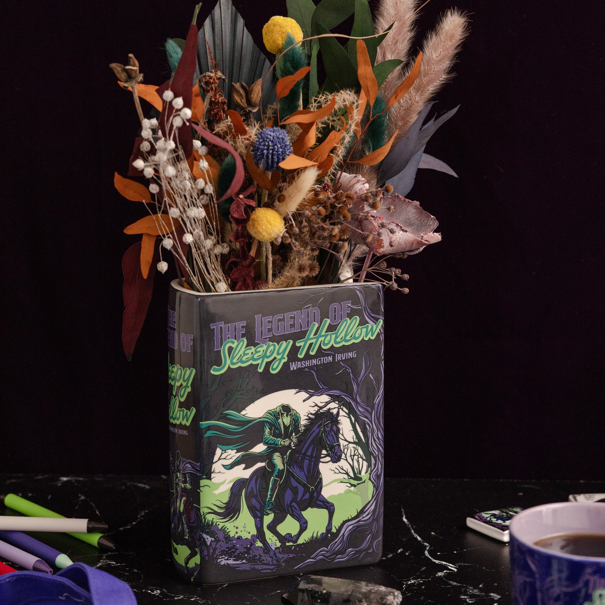 A decorative book-shaped vase featuring 'The Legend of Sleepy Hollow' artwork, filled with dried autumn flowers and foliage, set on a dark tabletop against a black background.