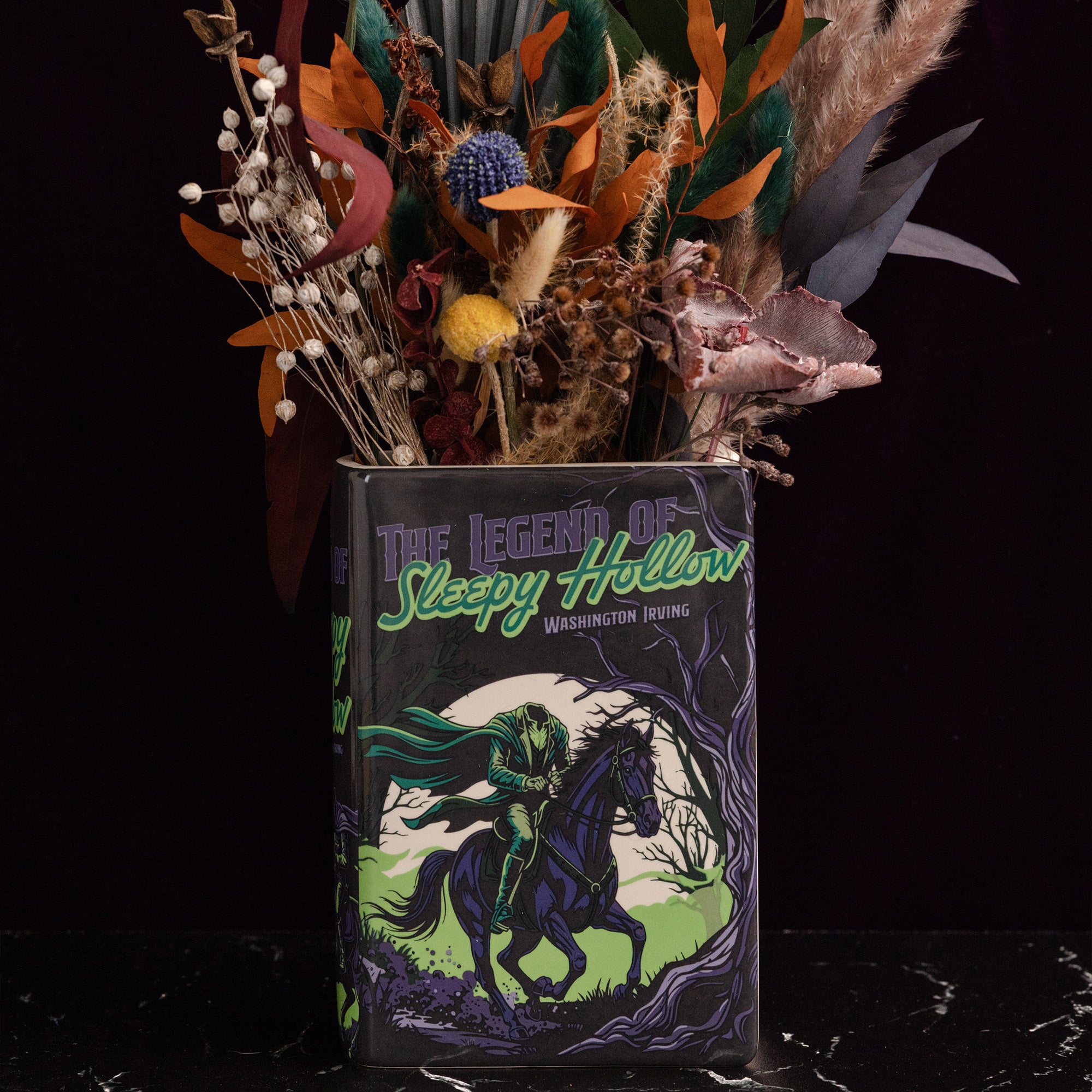 A decorative book-shaped vase featuring the cover art of 'The Legend of Sleepy Hollow' by Washington Irving, filled with dried autumn flowers and foliage against a dark background.