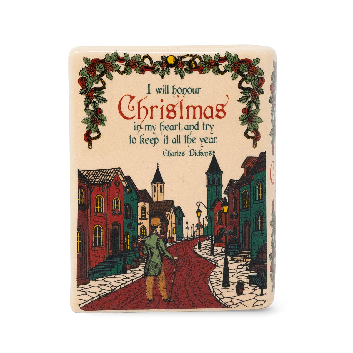 A rectangular ceramic Christmas candle featuring a vintage-style illustration of a cobblestone street with old buildings, a walking figure, and a green garland with red ornaments framing the text, 'I will honour Christmas in my heart, and try to keep it all the year.' by Charles Dickens.