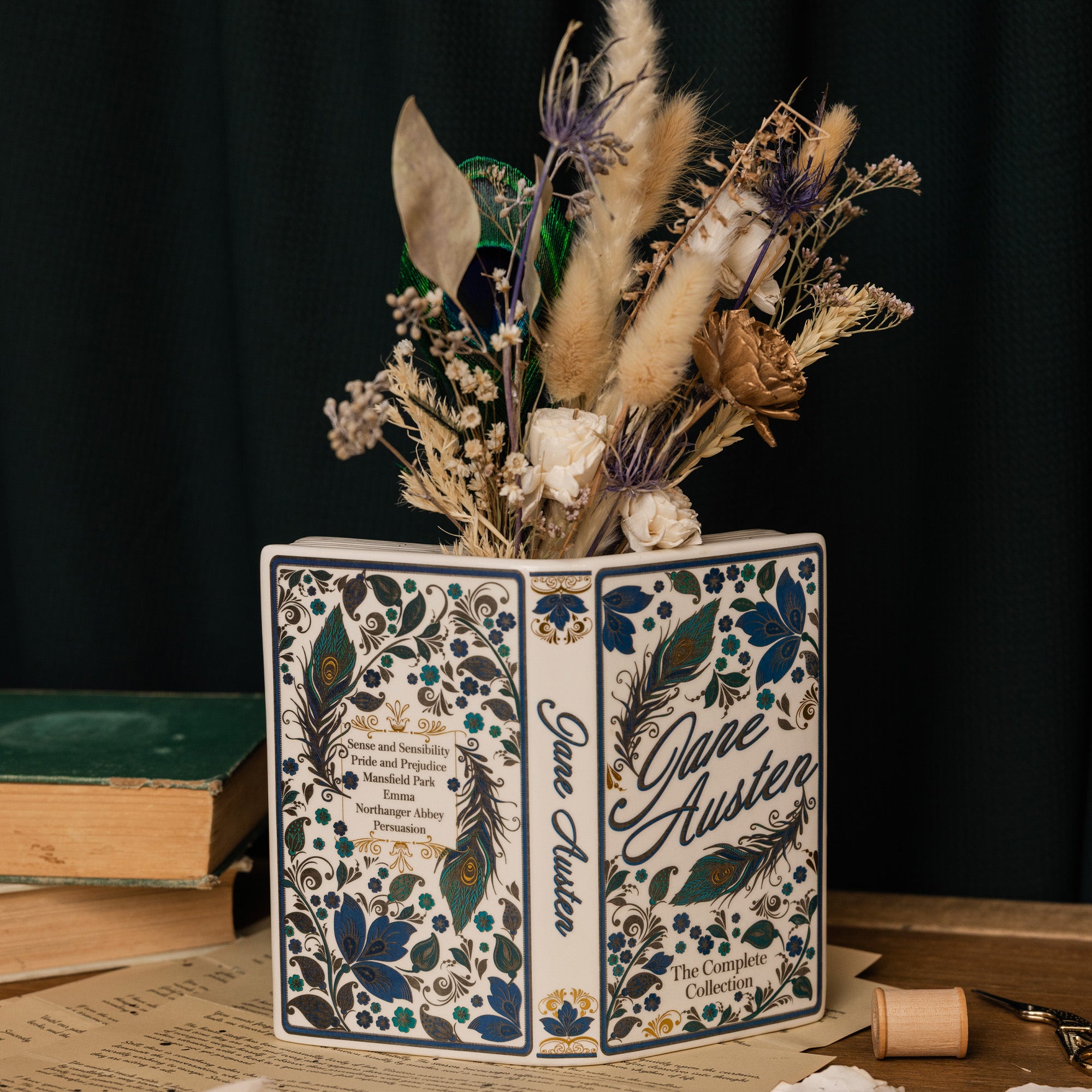 A decorative book-shaped vase featuring the title 'Jane Austen: The Complete Collection' with blue floral and peacock feather illustrations, filled with dried flowers and grasses, set on a tabletop with books in the background.