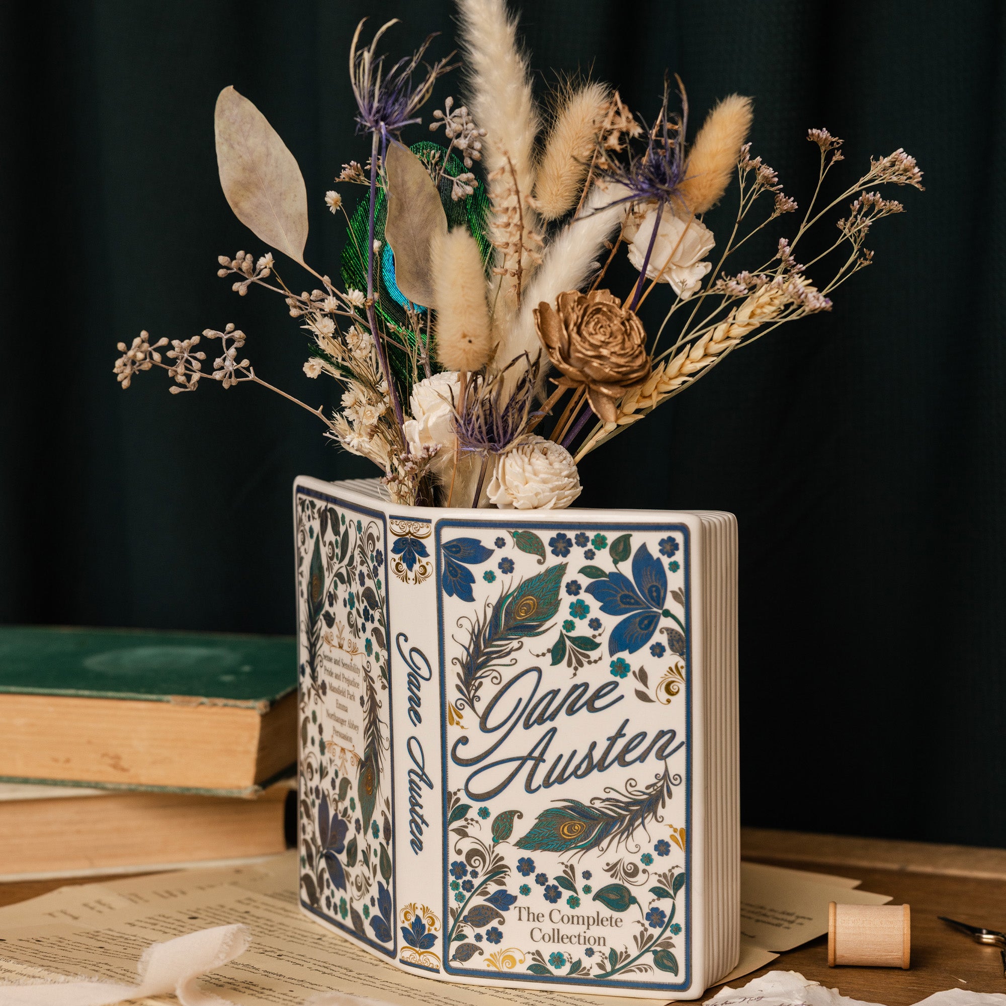 A decorative book-shaped vase featuring the title 'Jane Austen: The Complete Collection' with blue floral and peacock feather illustrations, filled with dried flowers and grasses, set on a tabletop with books in the background.